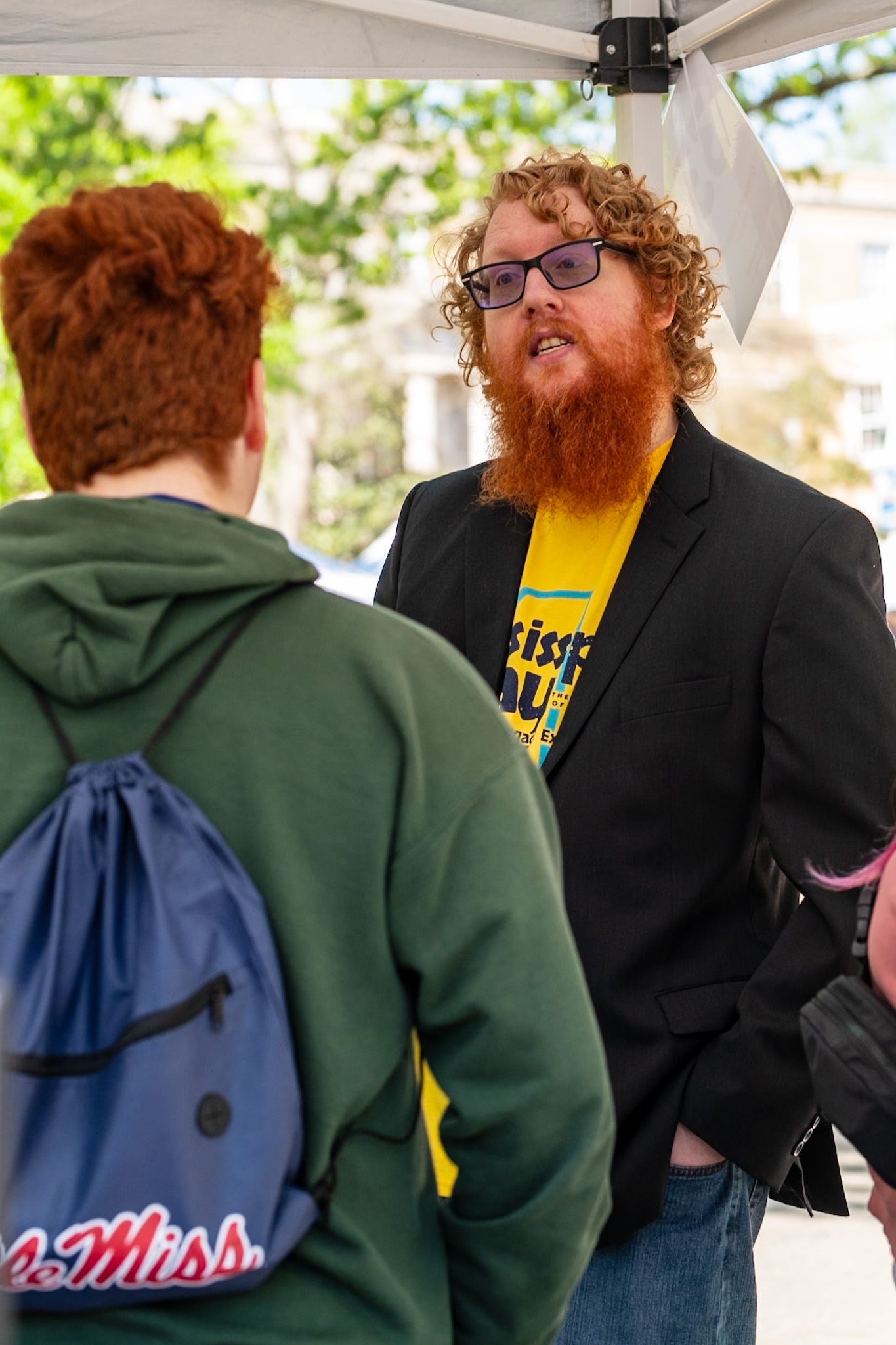 Two redheaded men conversing outdoors under a tent, one wearing a black blazer over a yellow shirt with a beard.
