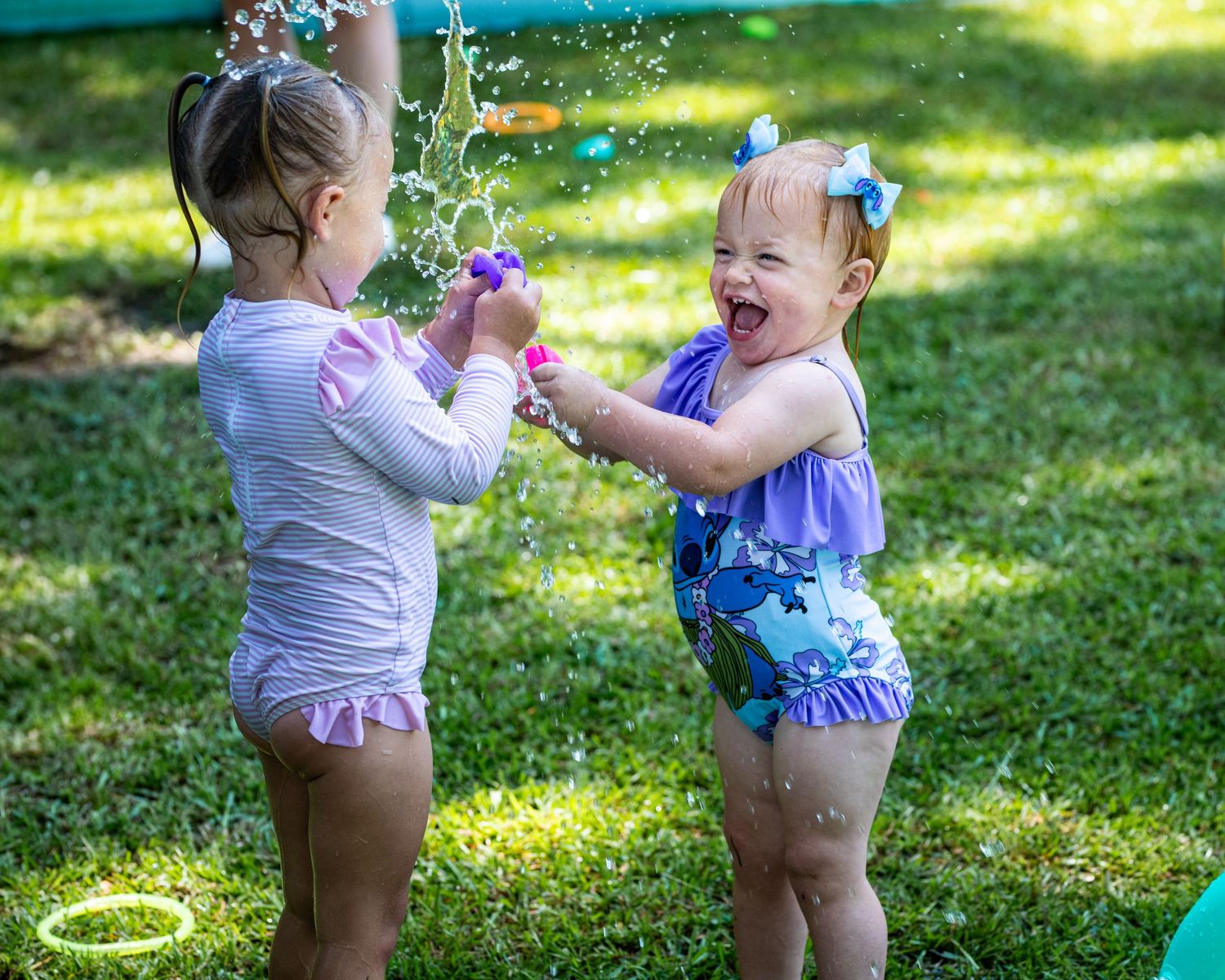Two toddler girls laughing and playing with water guns in swimsuits on a sunny green lawn.