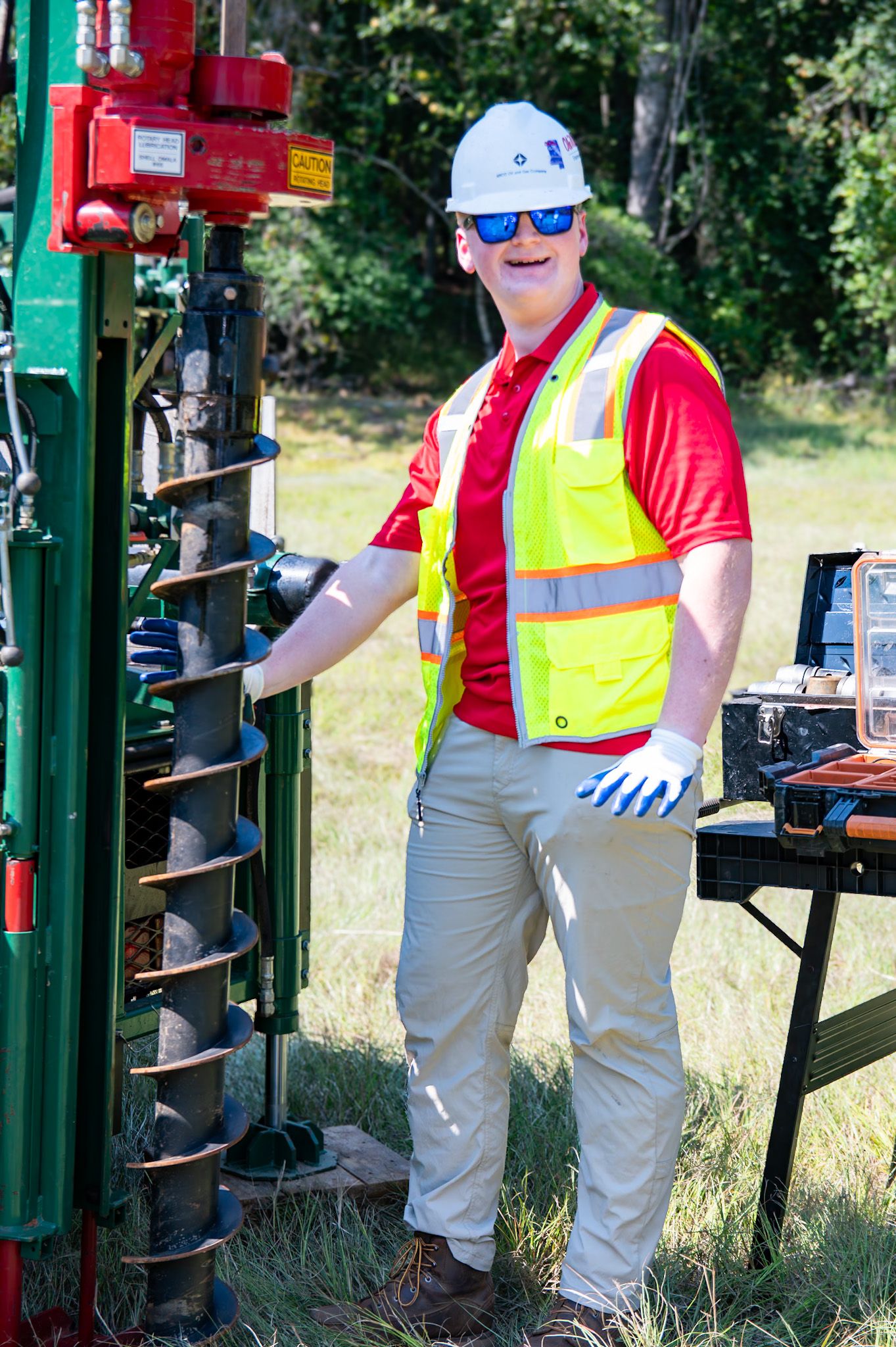 Worker in safety vest, hard hat, and sunglasses stands beside drilling equipment on a grassy construction site.