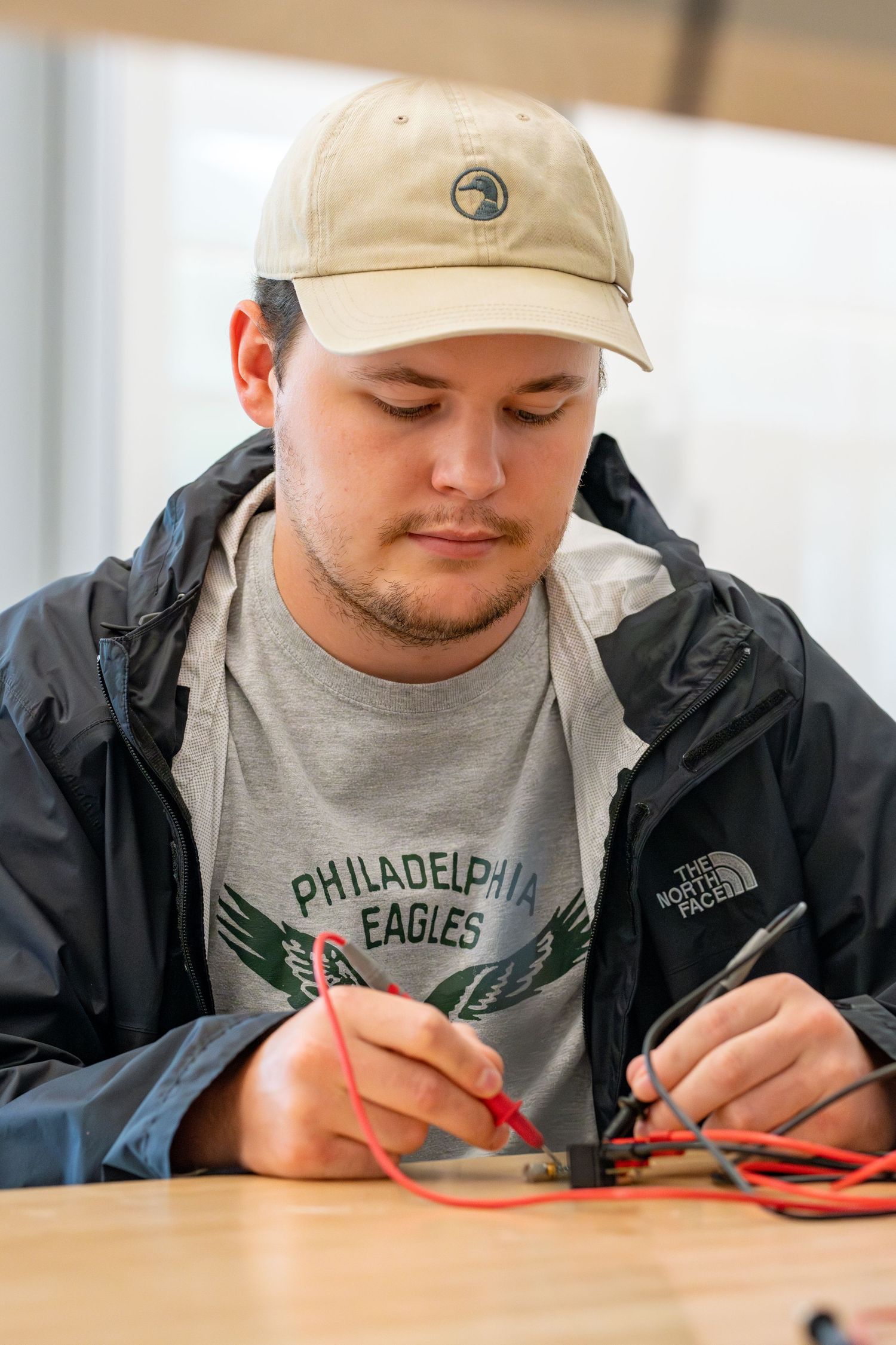 Young man in Philadelphia Eagles shirt and North Face jacket works with electrical wires at a wooden table.