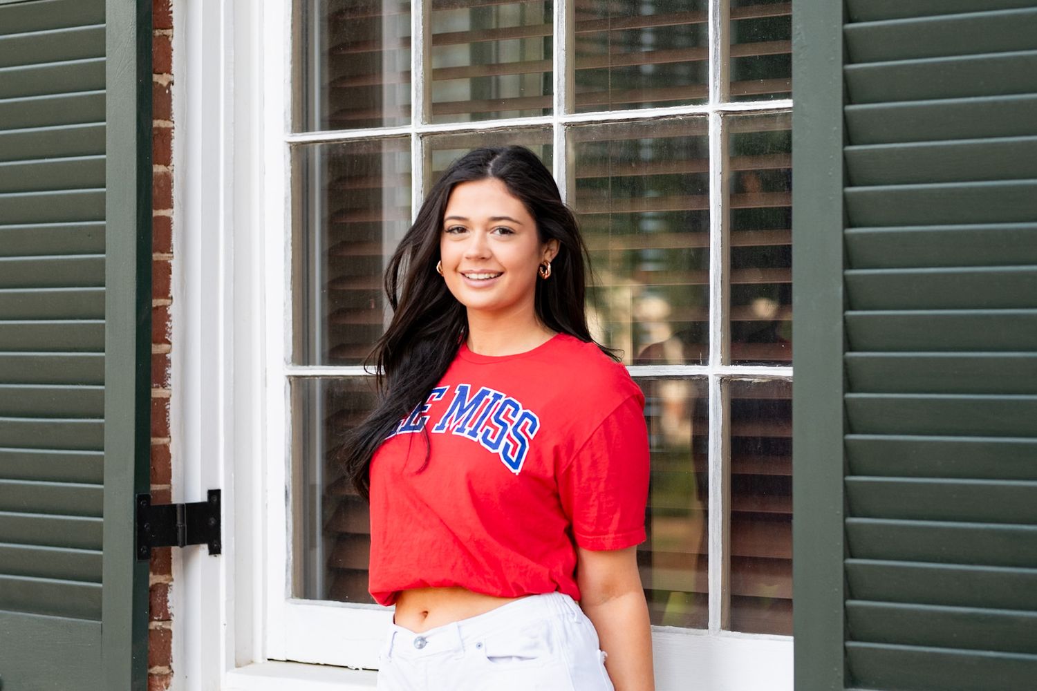 Young woman smiling in a red Ole Miss crop top, posing by a white window with green shutters.