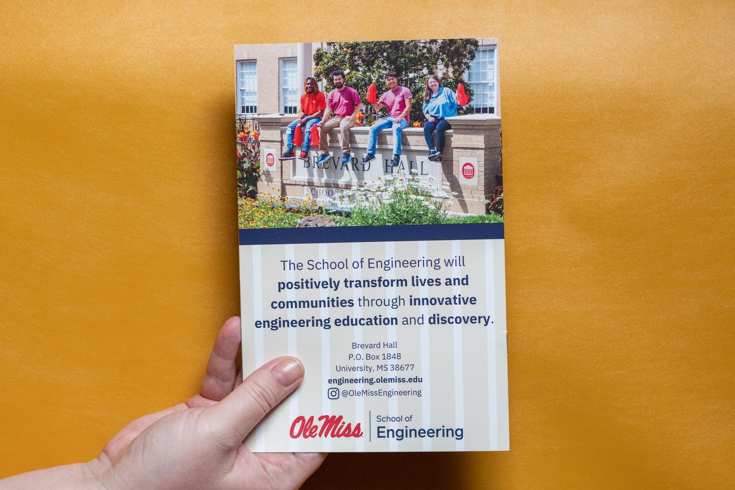 Hand holding Ole Miss School of Engineering brochure featuring students and mission statement on wooden background.