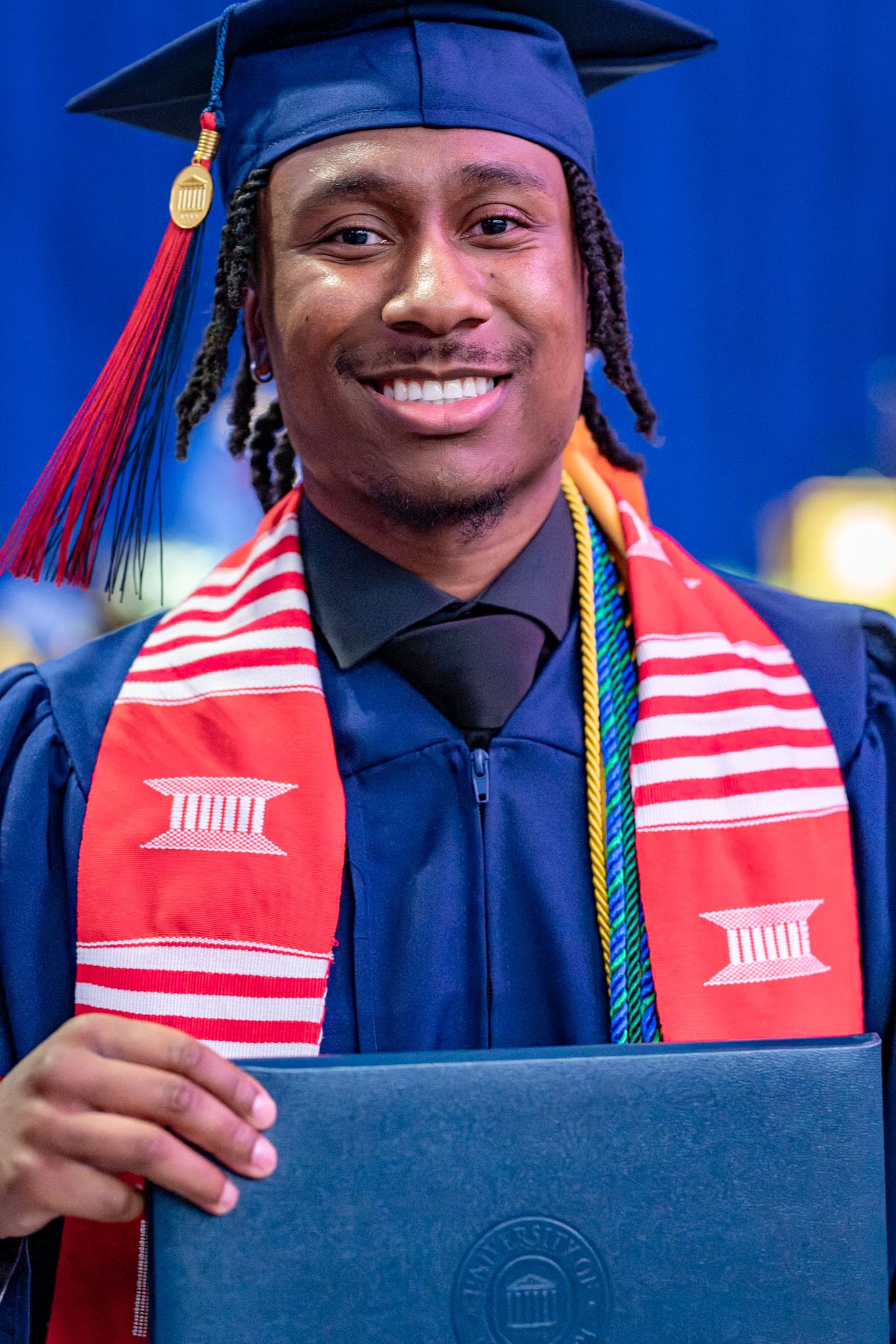 Graduate in blue cap and gown with red graduation stole and colorful cords holds diploma against blue background.