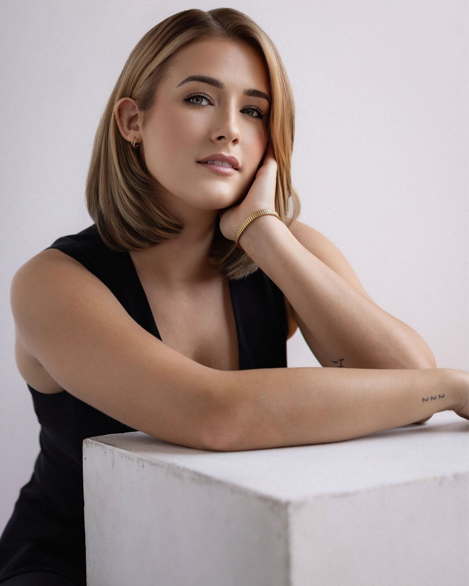 Studio portrait of person with shoulder-length brown hair named Mikiah Nicole Dixon wearin black sleeveless top while leaning on white cube.