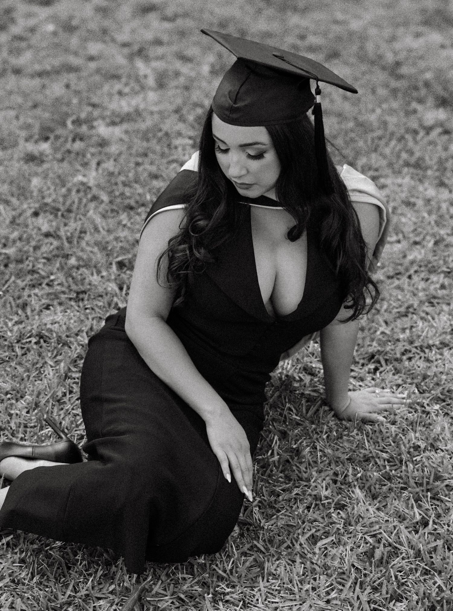 Graduate in black dress and cap poses on grass in black and white photo. Photo shot by Mikiah Nicole Dixon