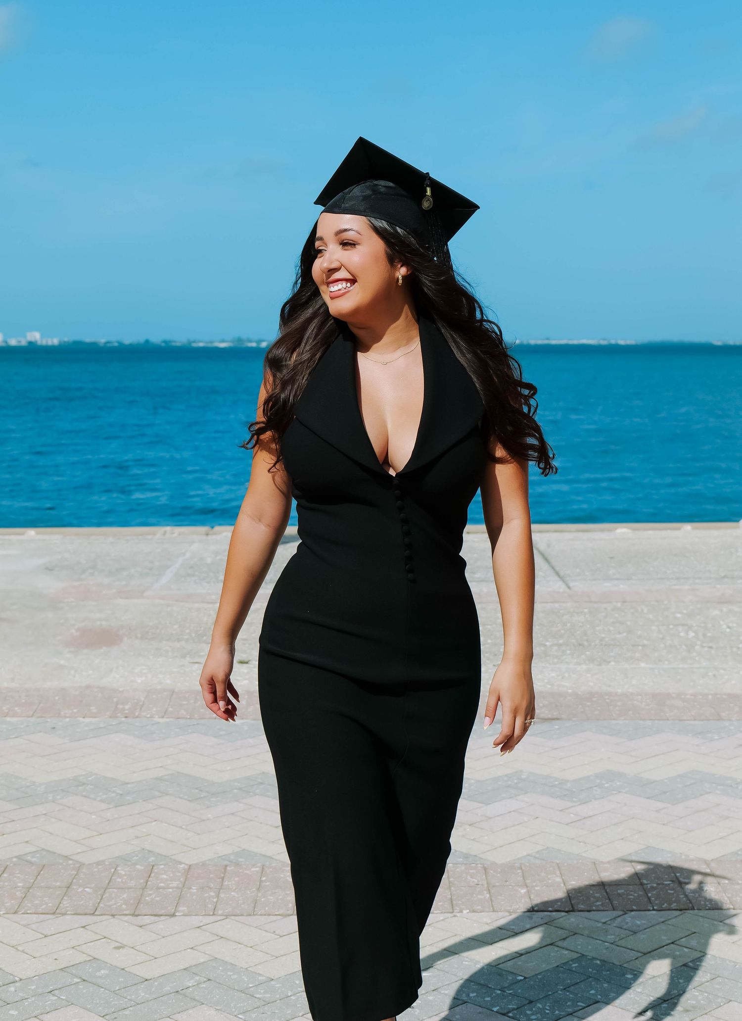 Graduate in black dress and cap smiles while walking along oceanfront boardwalk against bright blue water.Photo shot by Mikiah Nicole Dixon
