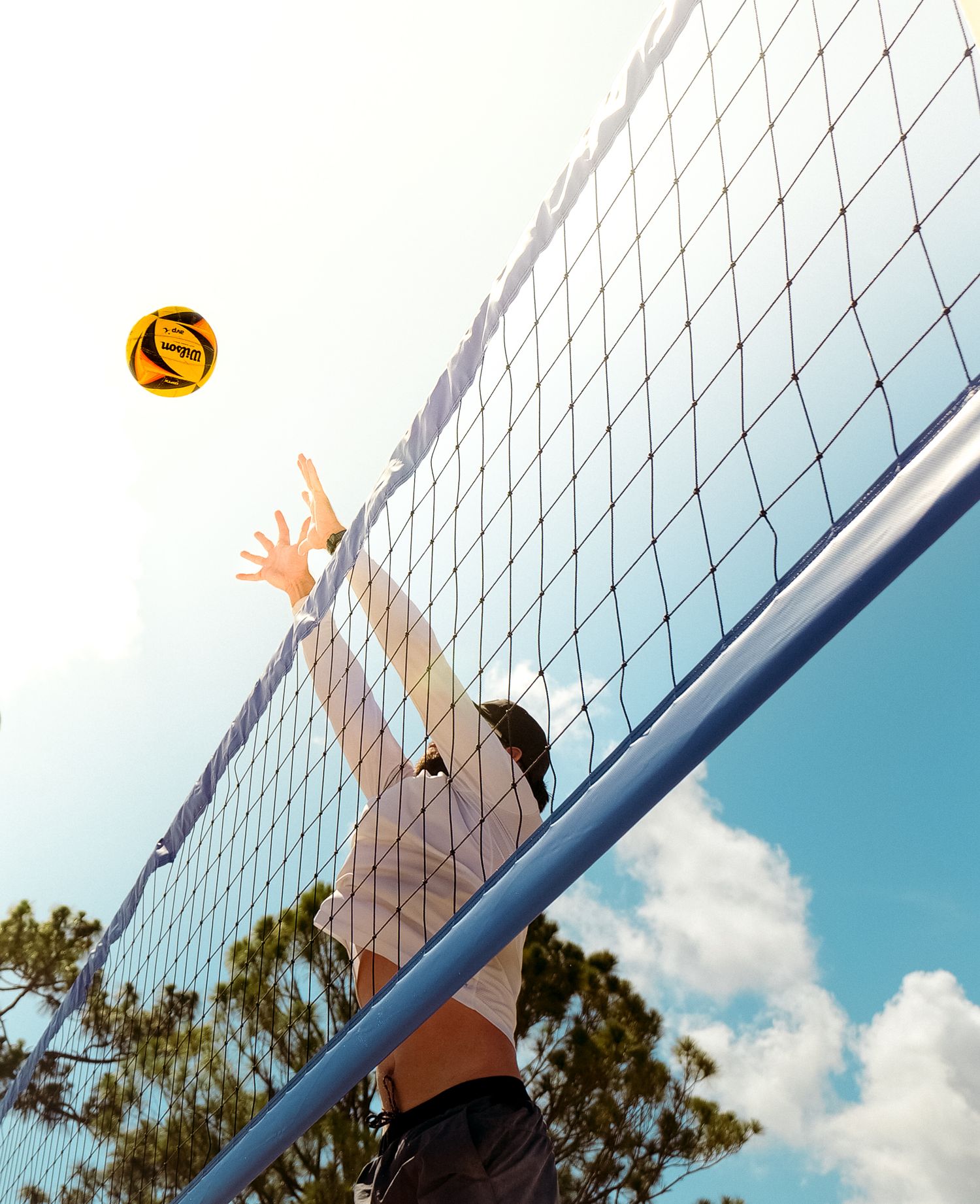 Beach volleyball player reaching up to block at the net against blue sky. Photo shot by Mikiah Nicole Dixon