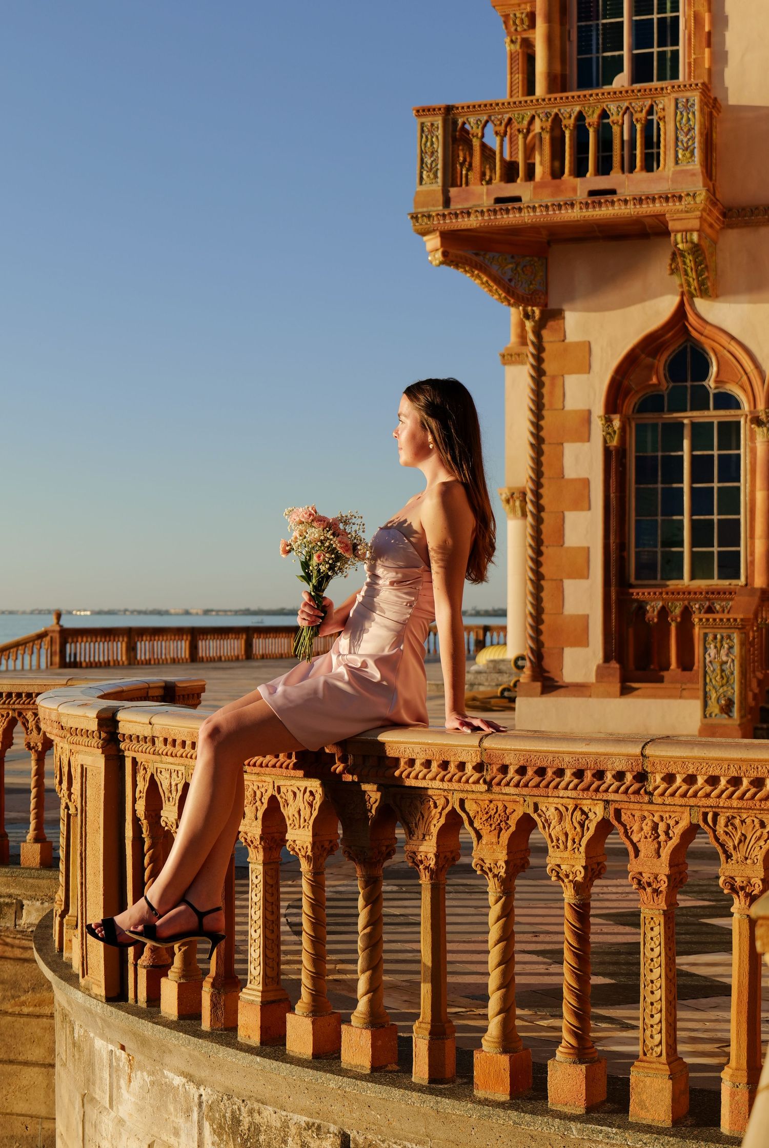 Woman in soft pink dress sits on decorative stone balcony of historic building. Photo shot by Mikiah Nicole Dixon