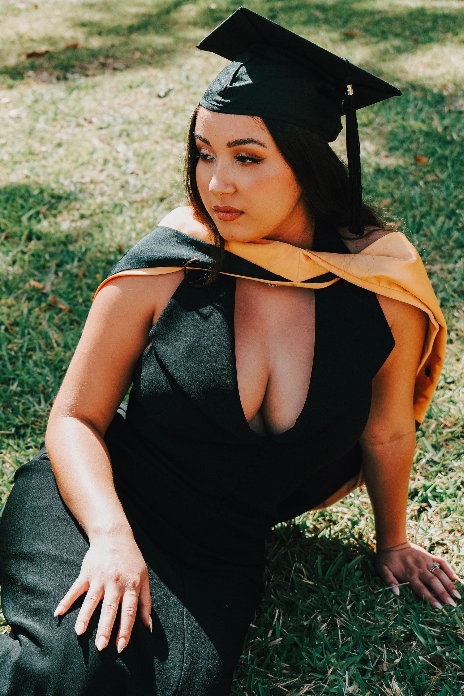 Graduate wearing cap and gown poses outdoors on sunny grass. Photo shot by Mikiah Nicole Dixon