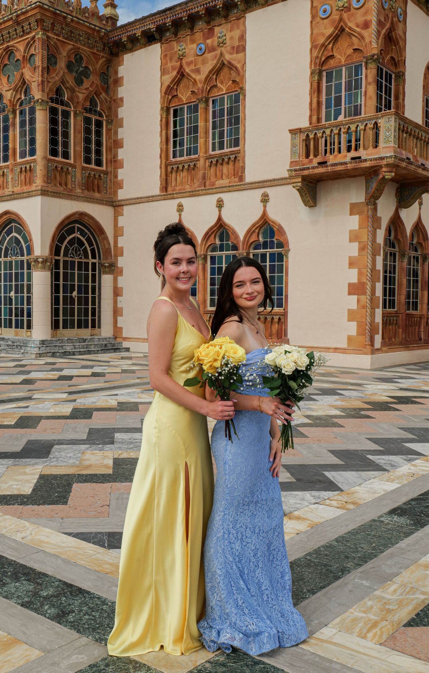 Two women in yellow and blue formal gowns posing at ornate Mediterranean-style building. Photo shot by Mikiah Nicole Dixon