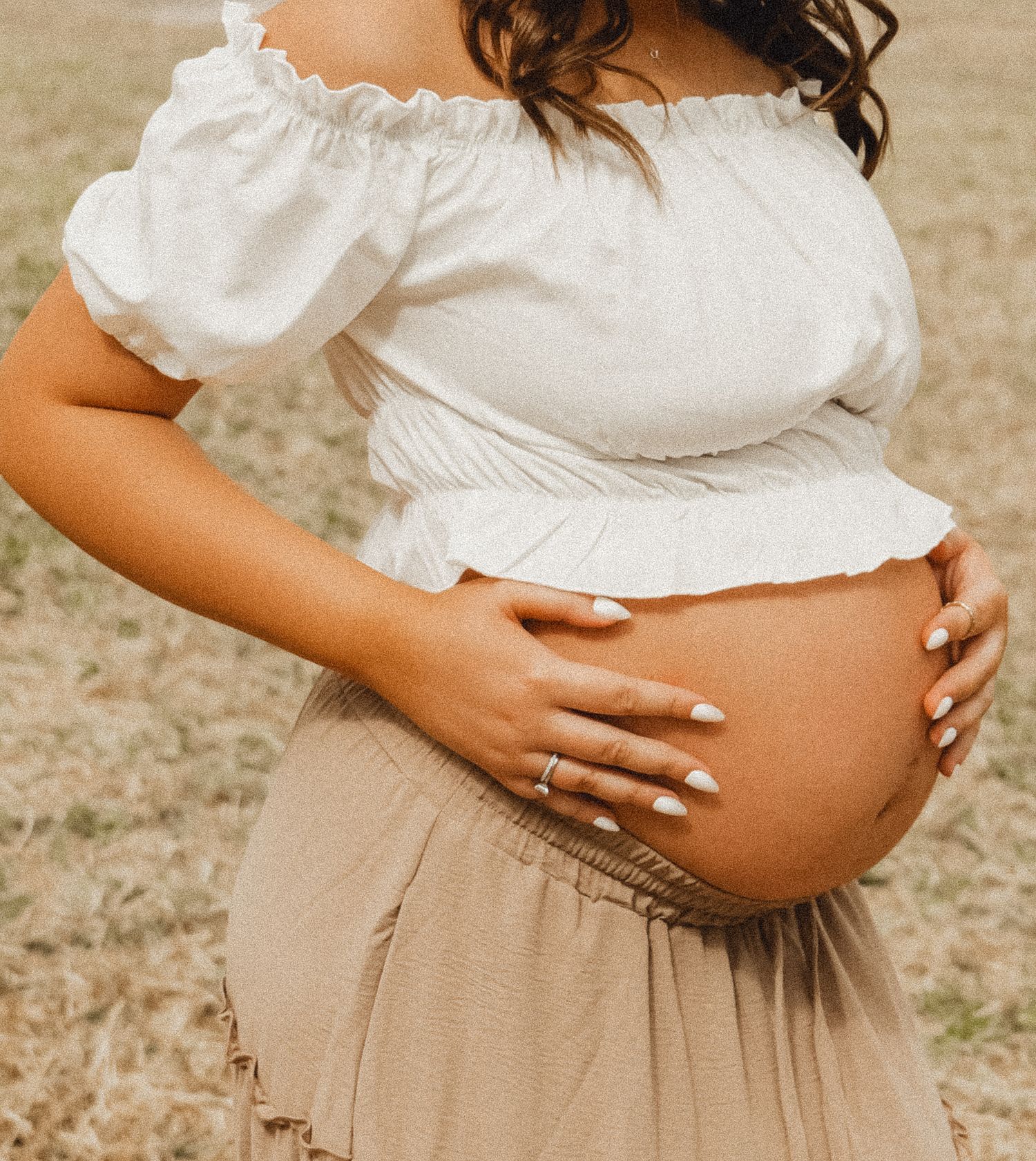 A close-up photo of a pregnant belly with hands wearing a white peasant top and beige skirt. Photo shot by Mikiah Nicole Dixon