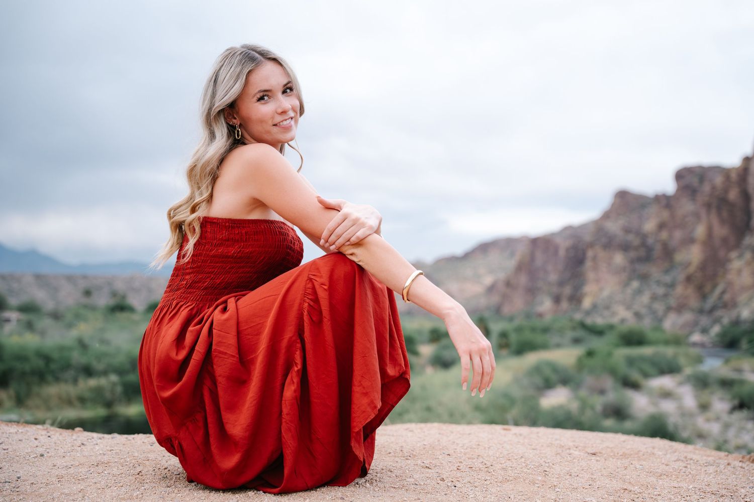 Des Moines Iowa senior portrait photographer – joyful young woman in flowing red dress sitting on rocks with mountain backdrop, golden hour.