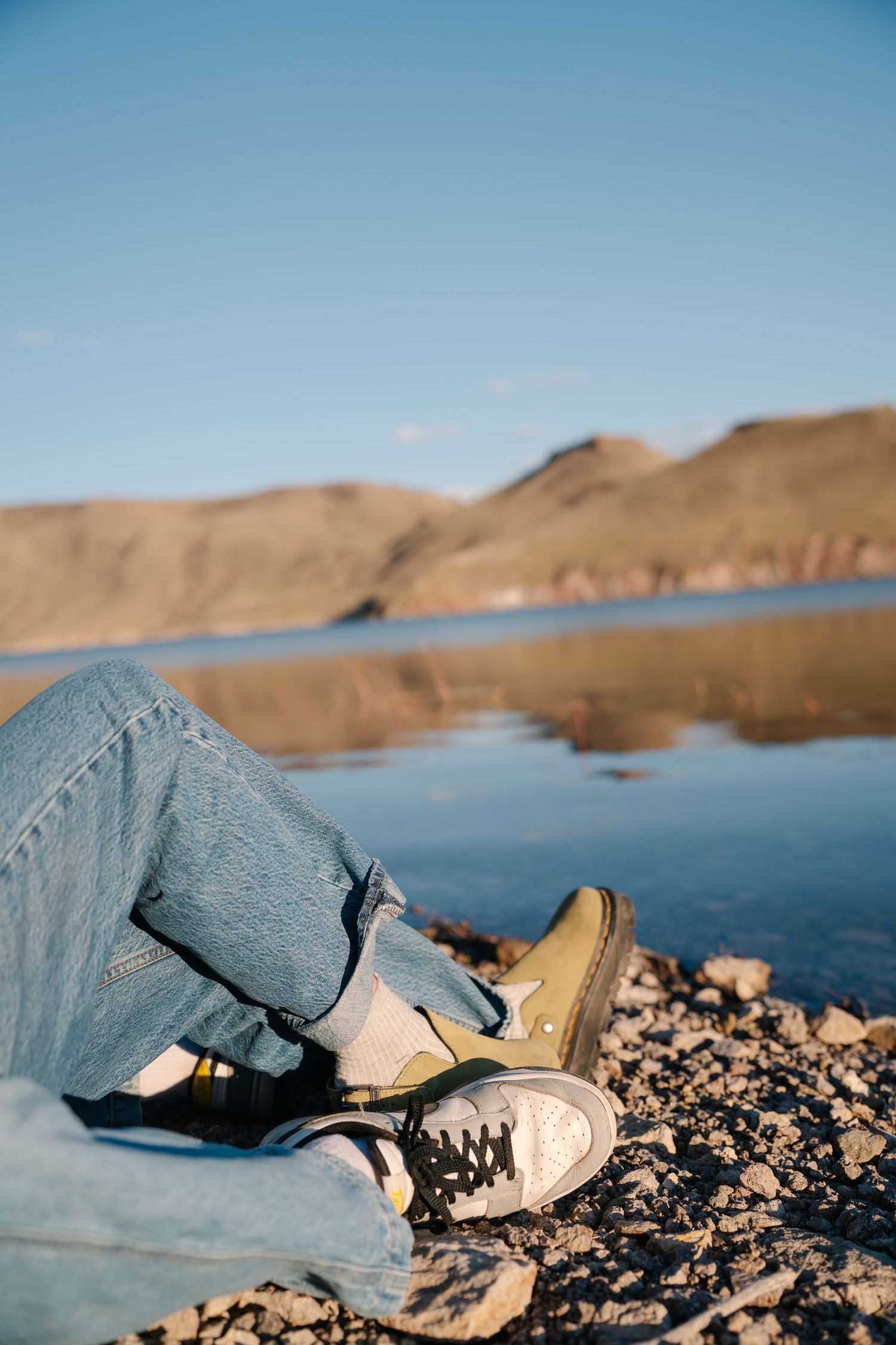 Des Moines Iowa lake engagement photography – casual couple sitting on rocky shore by blue water, relaxed jeans look with scenic view