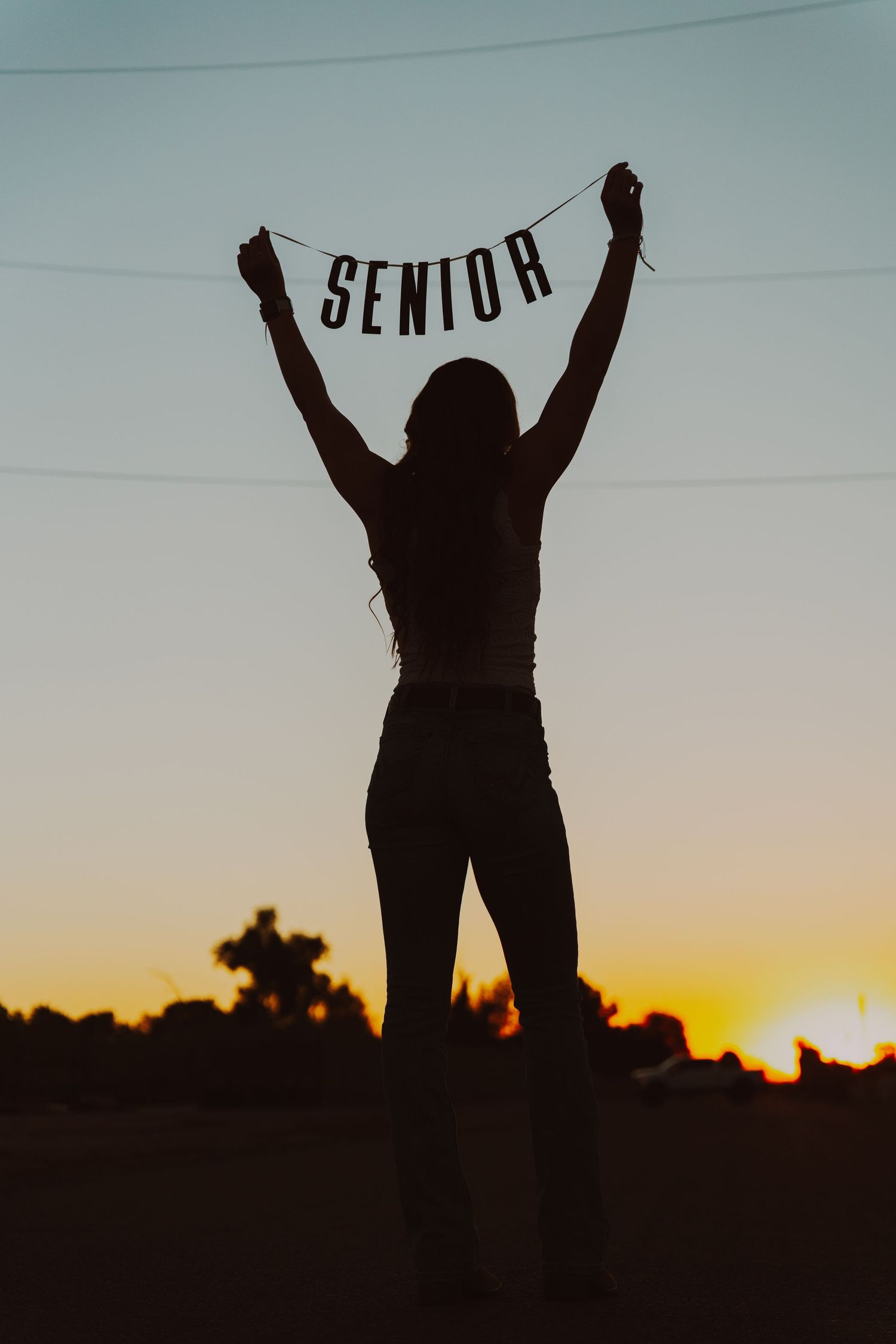 Des Moines Iowa senior portrait photographer – empowering silhouette of girl with arms raised at sunset, 'SENIOR' overlay in dramatic sky.