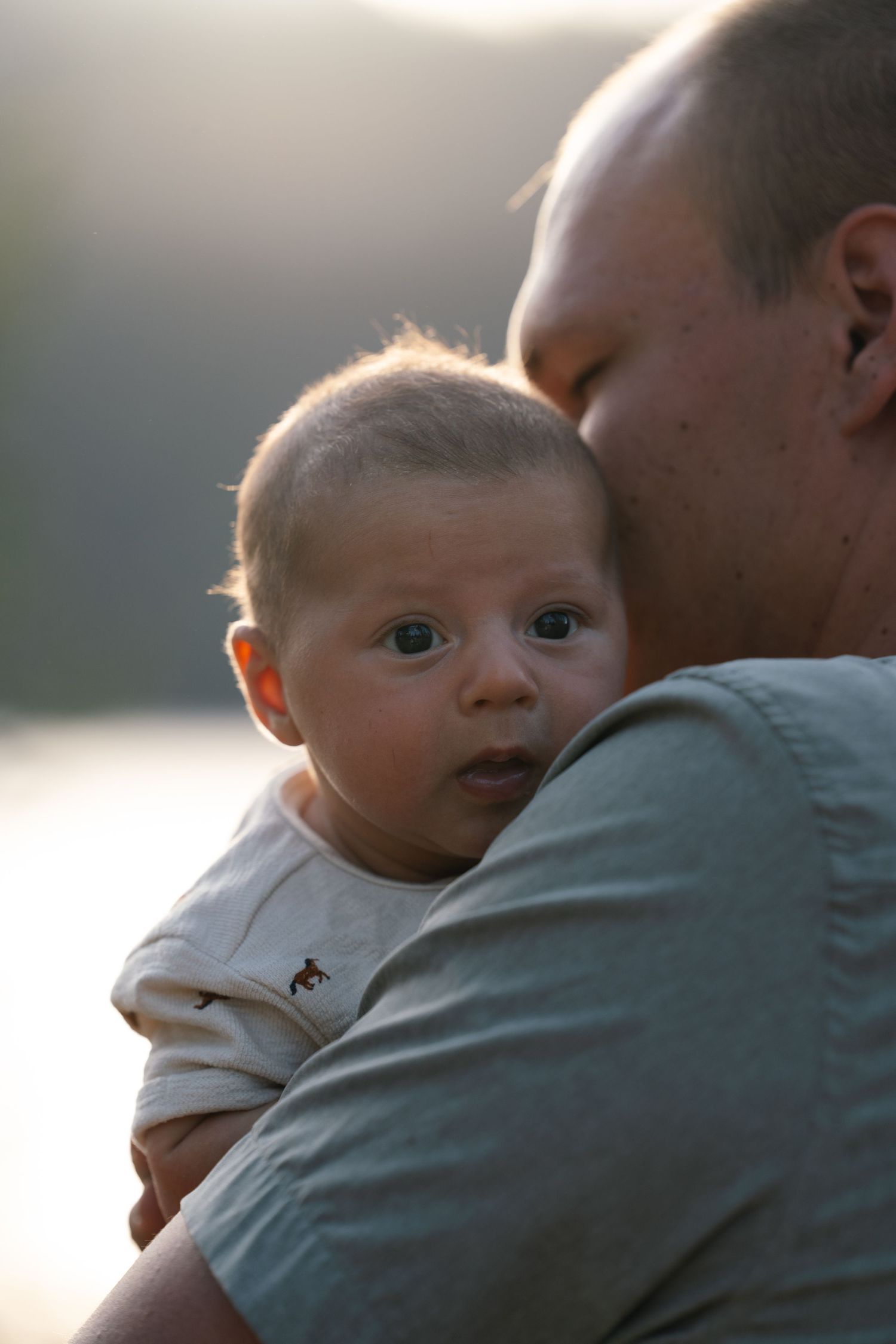 A young father holding his newborn son who is looking over the dad's shoulder at the camera in the golden hours.