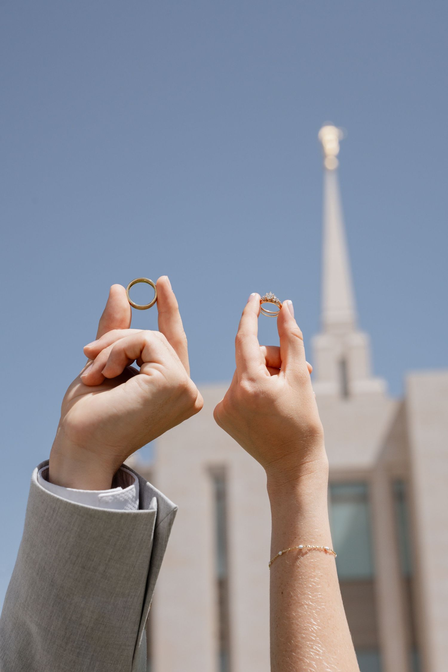 Des Moines wedding photography detail: bride and groom holding up wedding rings in front of modern temple architecture under summer blue sky