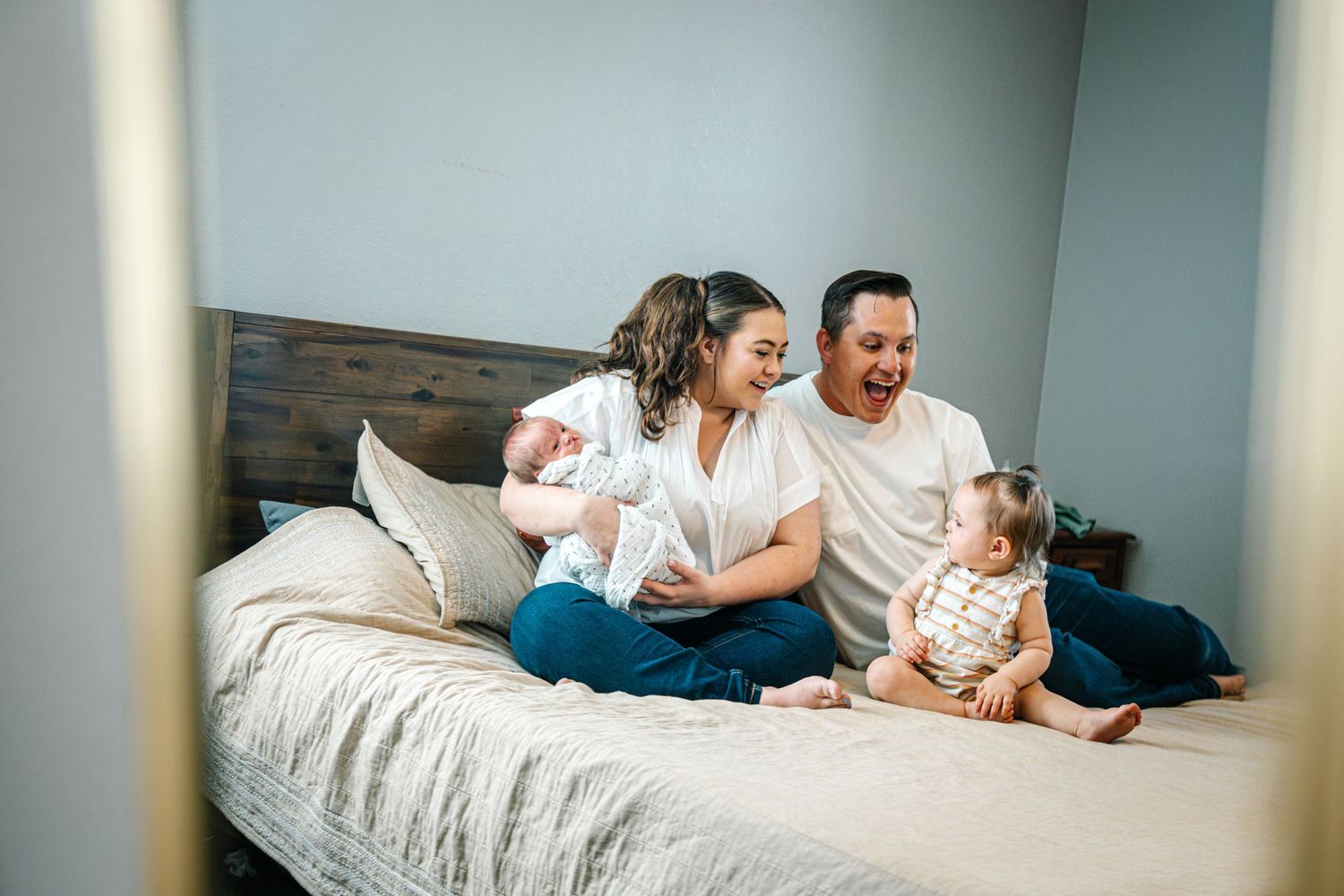 Des Moines Iowa newborn family photographer – tender home portrait of parents with newborn and sibling on bed, soft window light and loving.