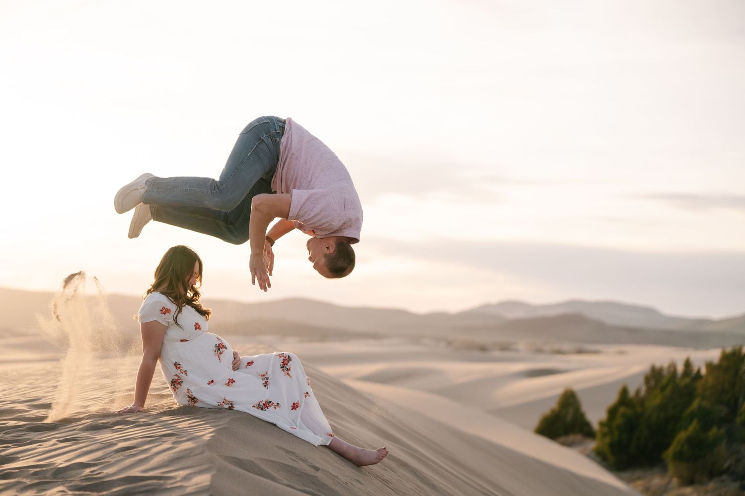 Des Moines Iowa maternity photographer – playful pregnant woman in white dress doing backflip on sand dunes at golden hour, joyful desert po