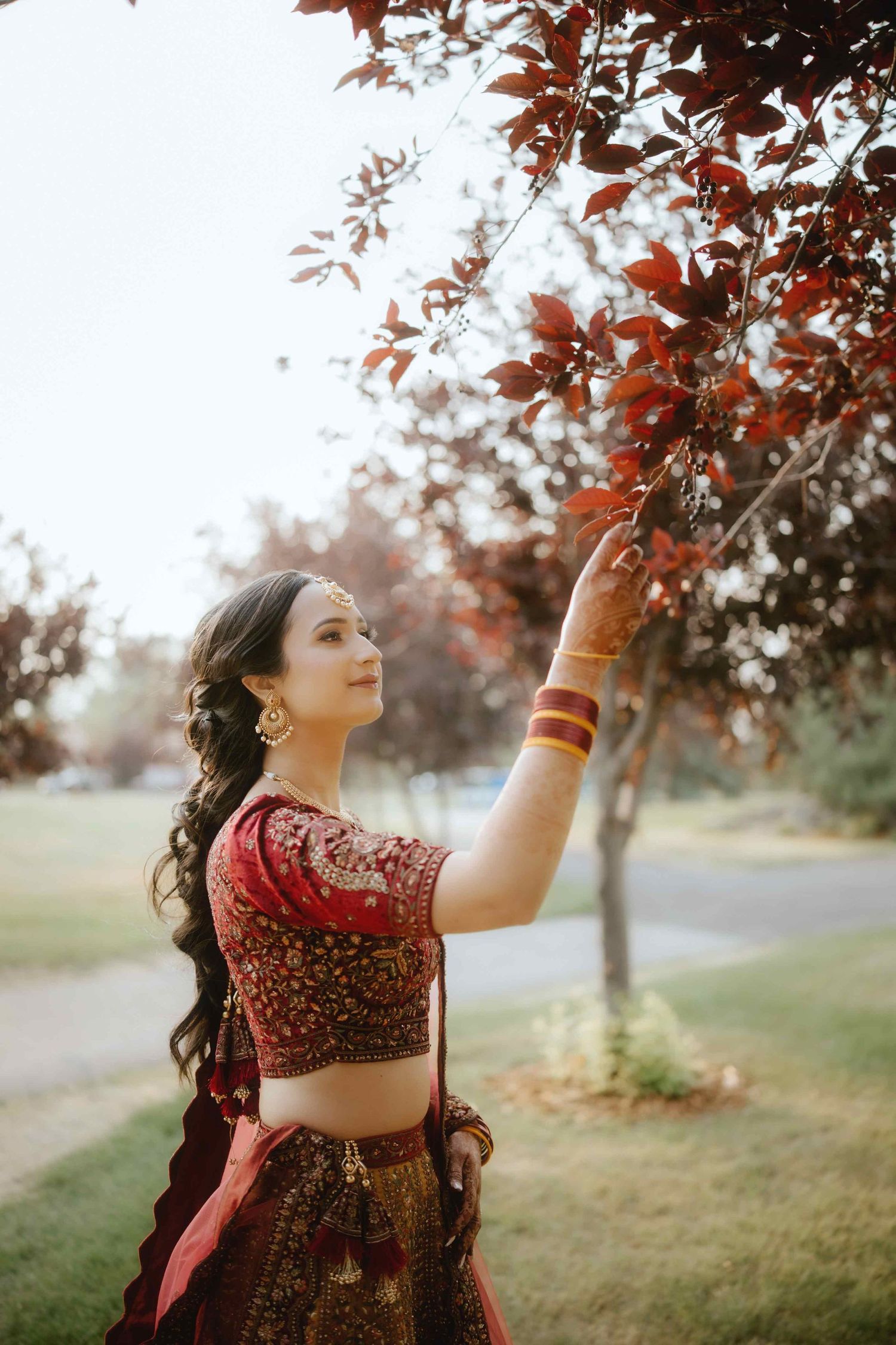 Traditional Indian bride in a deep red lehenga poses gracefully under autumn trees in an outdoor portrait session.
Jaago Day