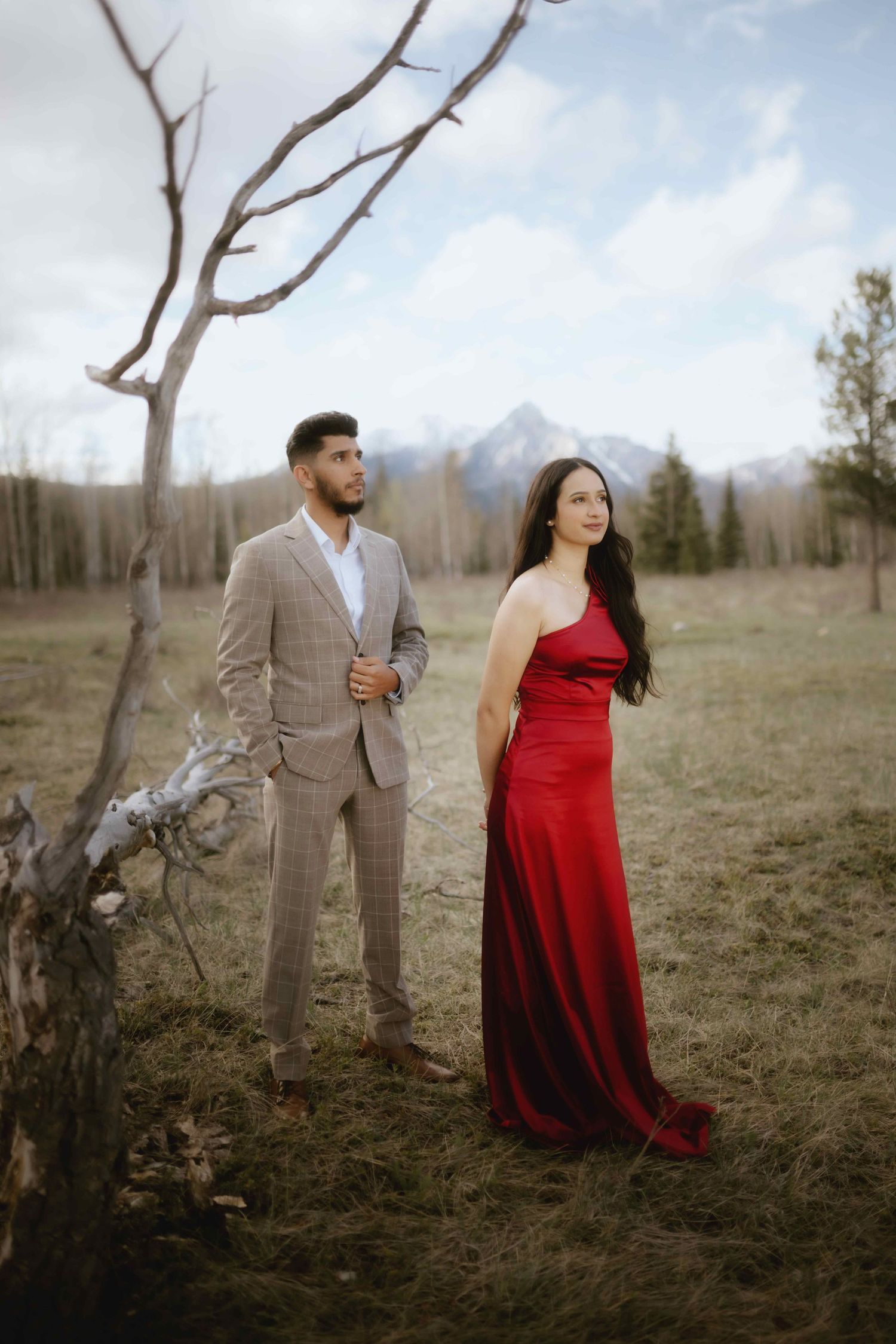 A couple wearing red and tan poses together in a field with mountains and bare trees in the background.
Banff Couple Shoot