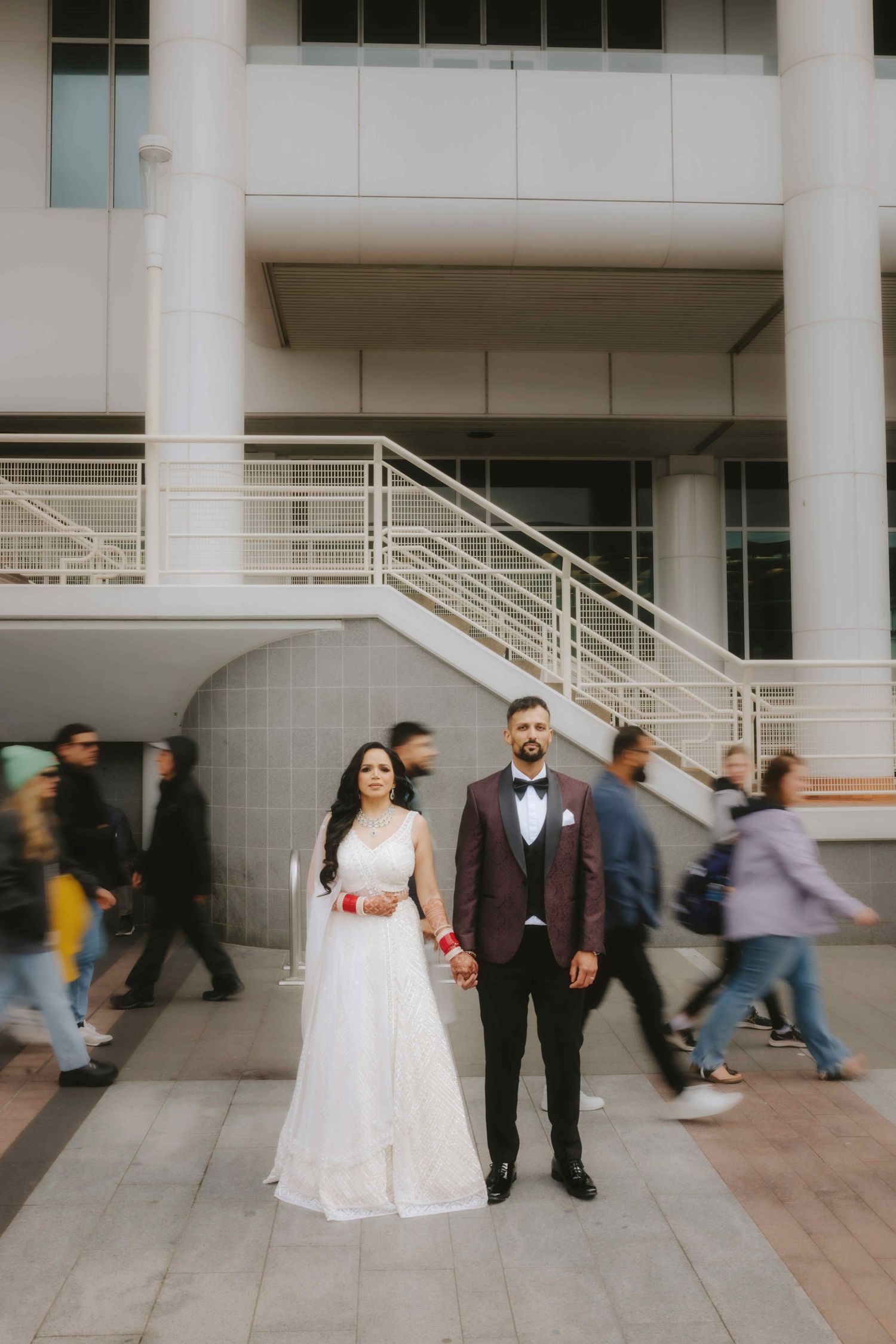A wedding couple poses in formal attire as people walk past a modern building entrance.
Reception photography