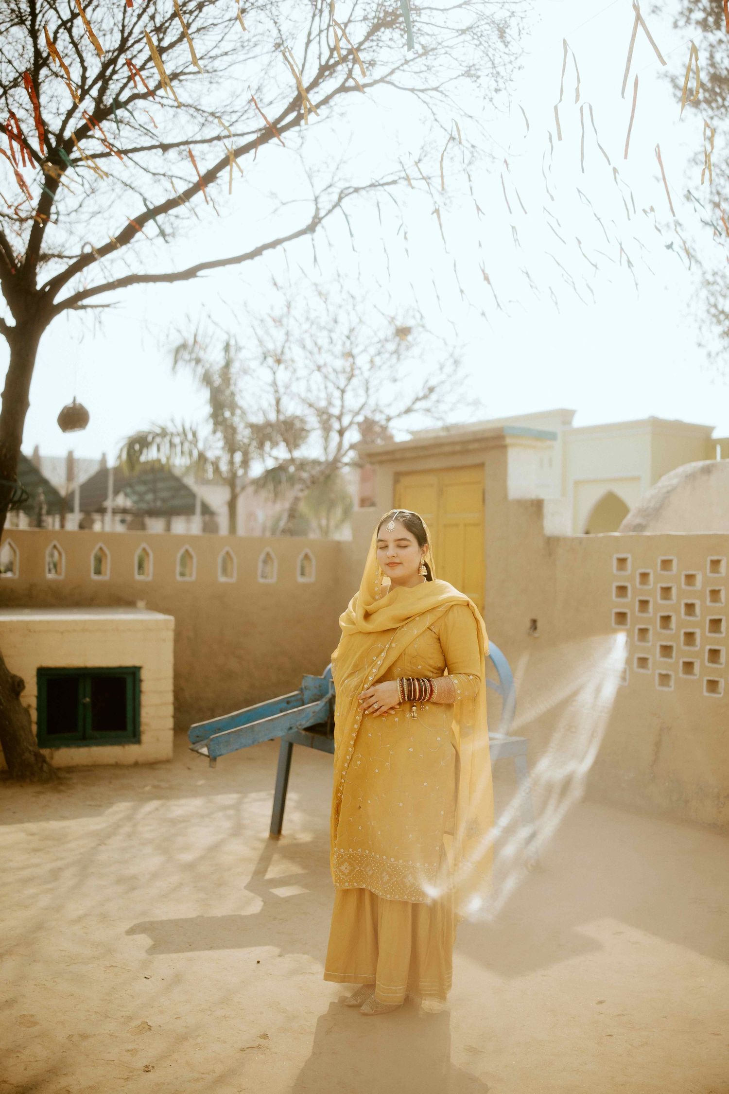 Person in yellow traditional outfit standing against adobe wall in sunlight.
Sikh Bride