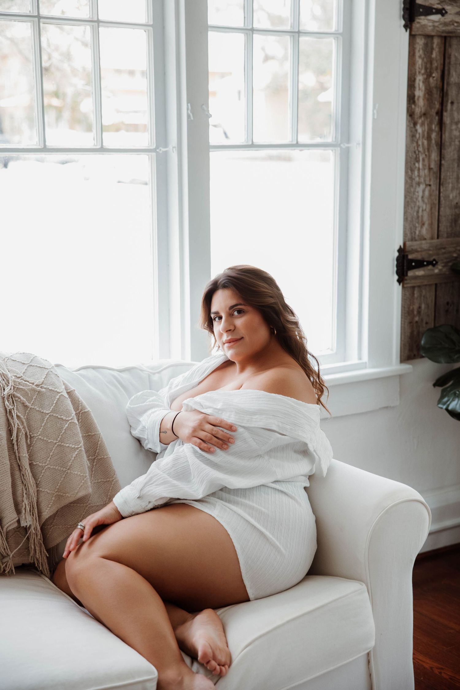 Women wearing white oversized sweater seated beside a window durning a boudoir session