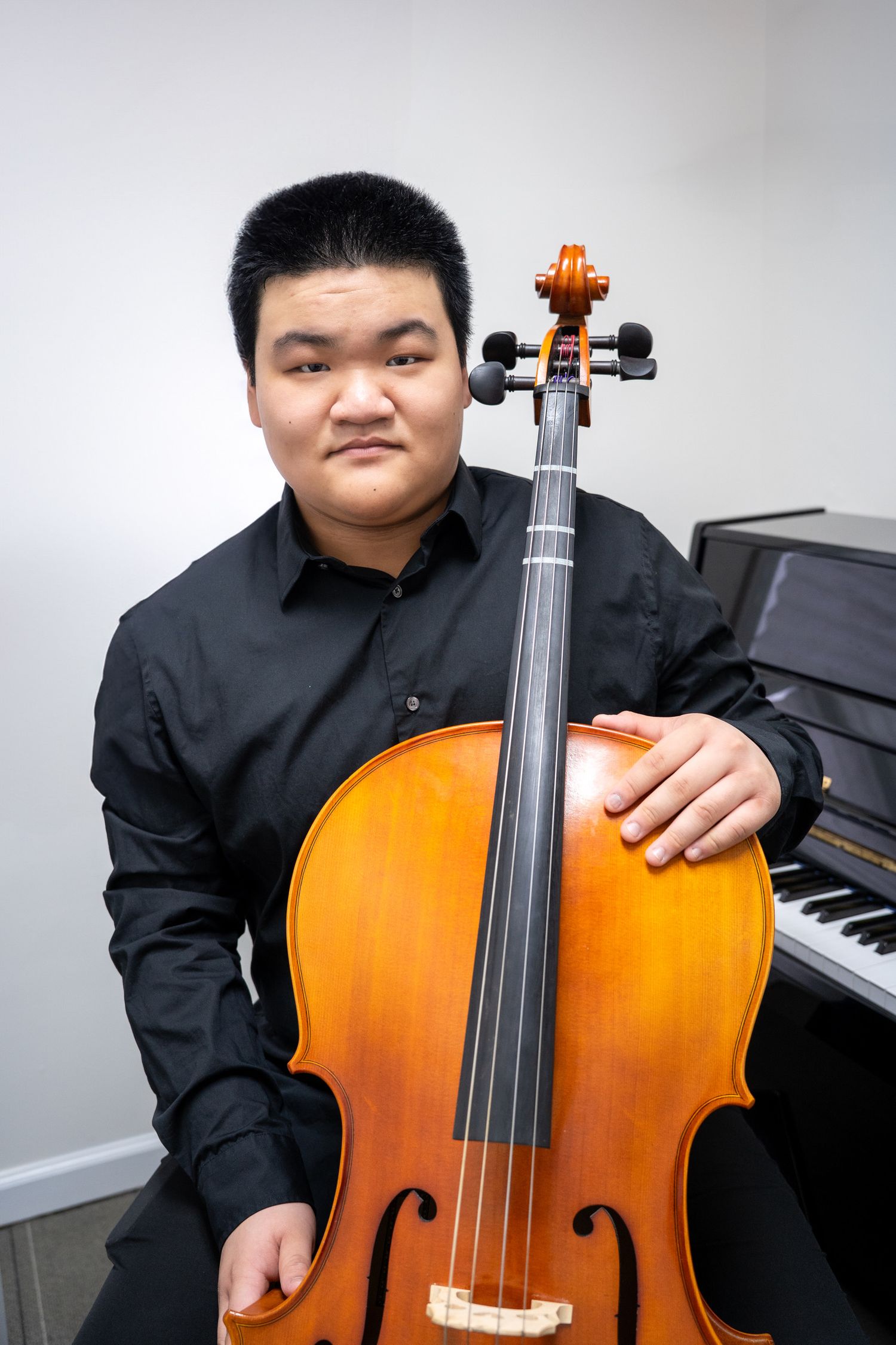 Portrait of a young man in a black shirt holding his cello in front of a piano.