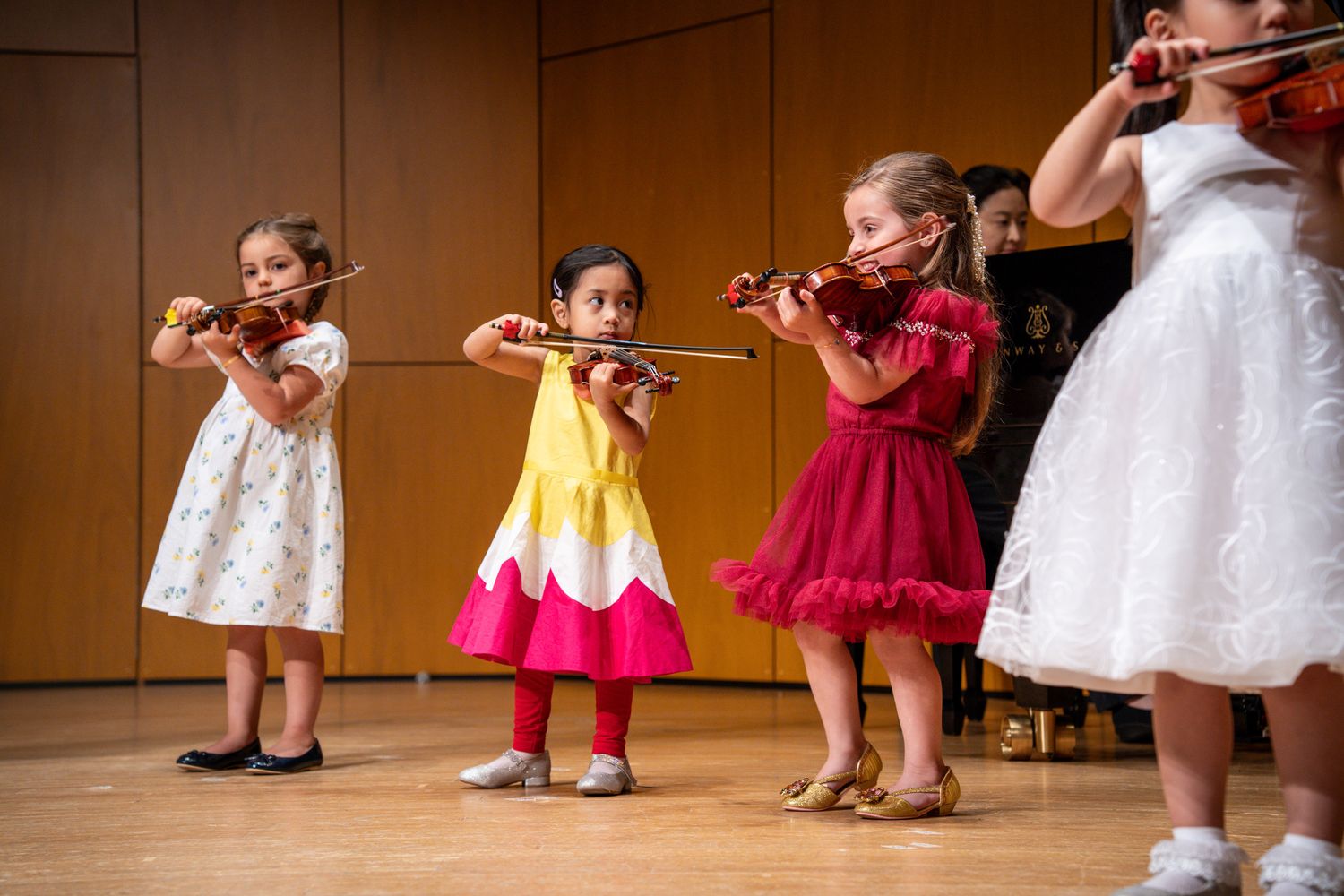 Four young girls in colorful dresses performing on violins on stage.