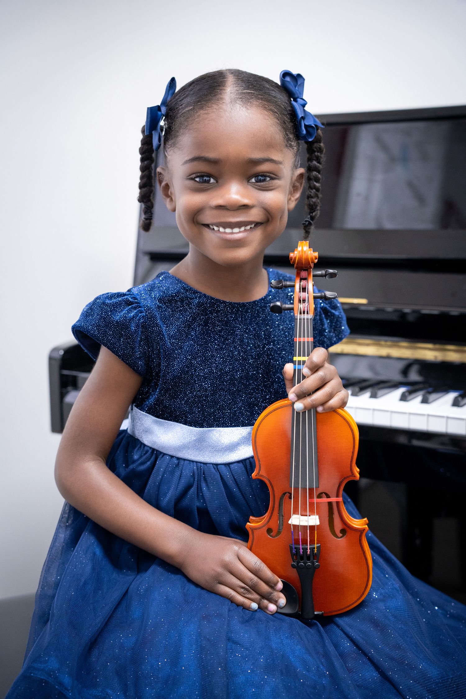 Portrait of a young girl in a blue dress holding her violin and smiling in front of a piano.