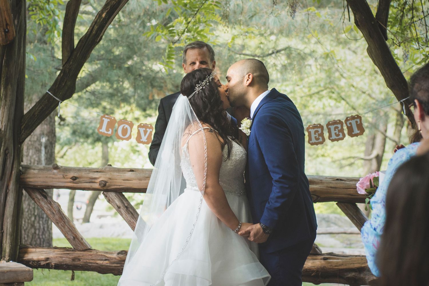 Bride and groom sharing a kiss under trees at an outdoor wedding ceremony.