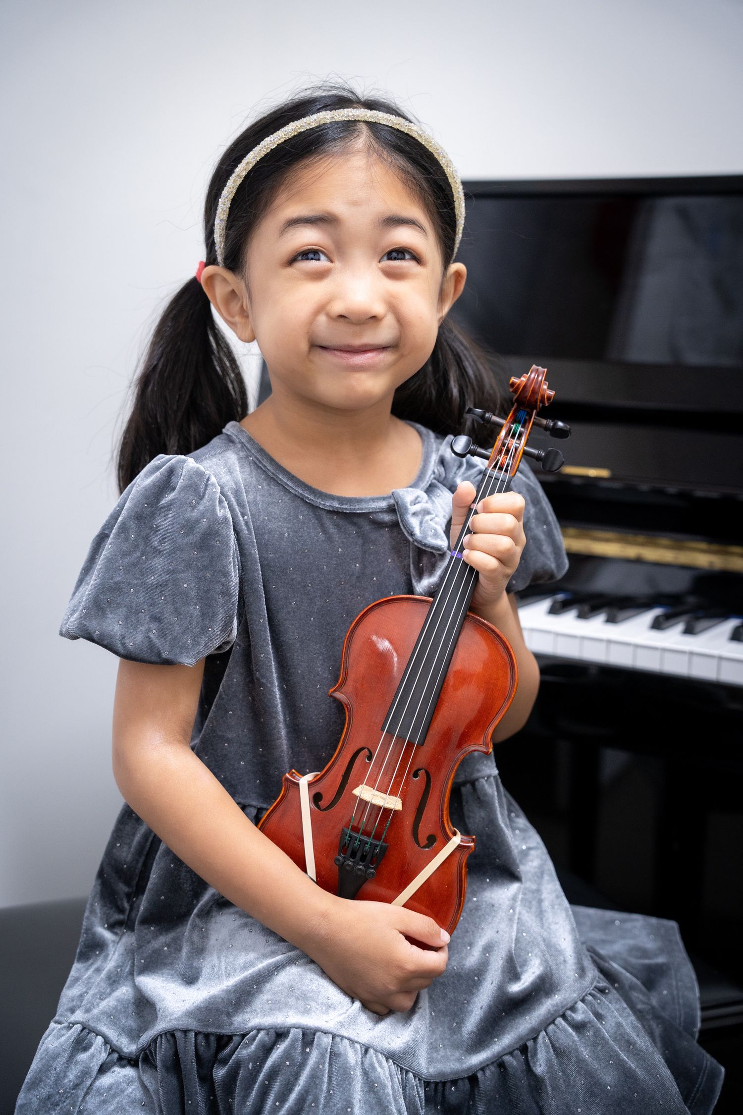 Portrait of a young girl in a gray dress holding her violin in front of a piano.