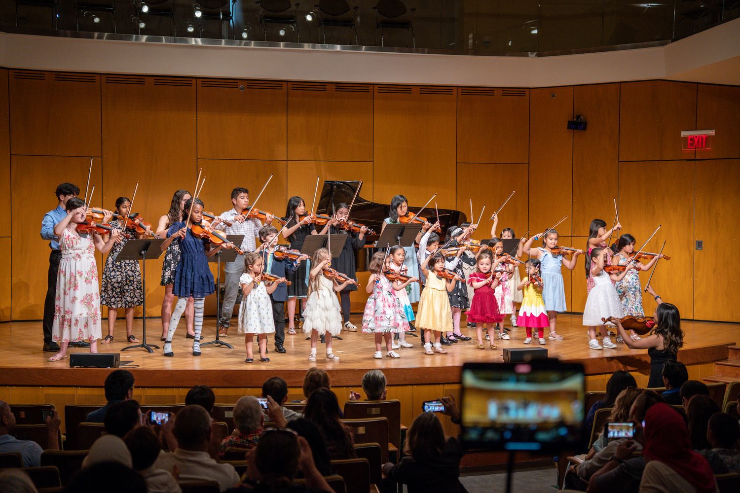 Large group of children performing a violin ensemble for an audience in a concert hall.