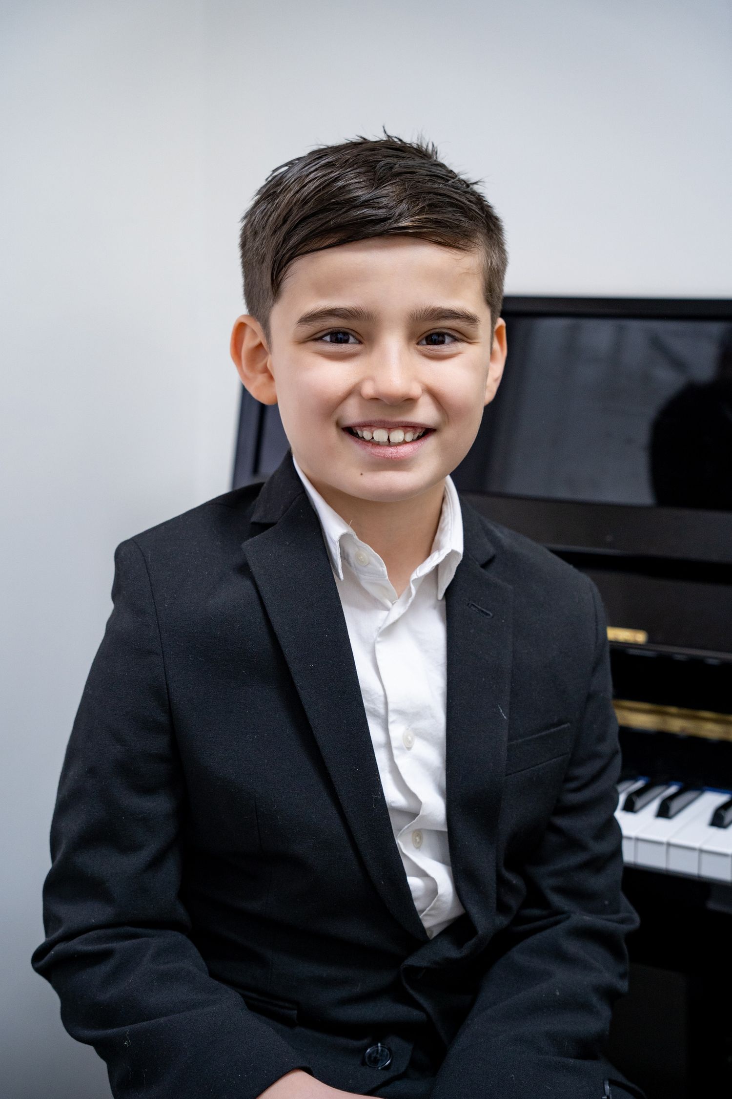 Portrait of a young boy in a black suit holding his violin in front of a piano.