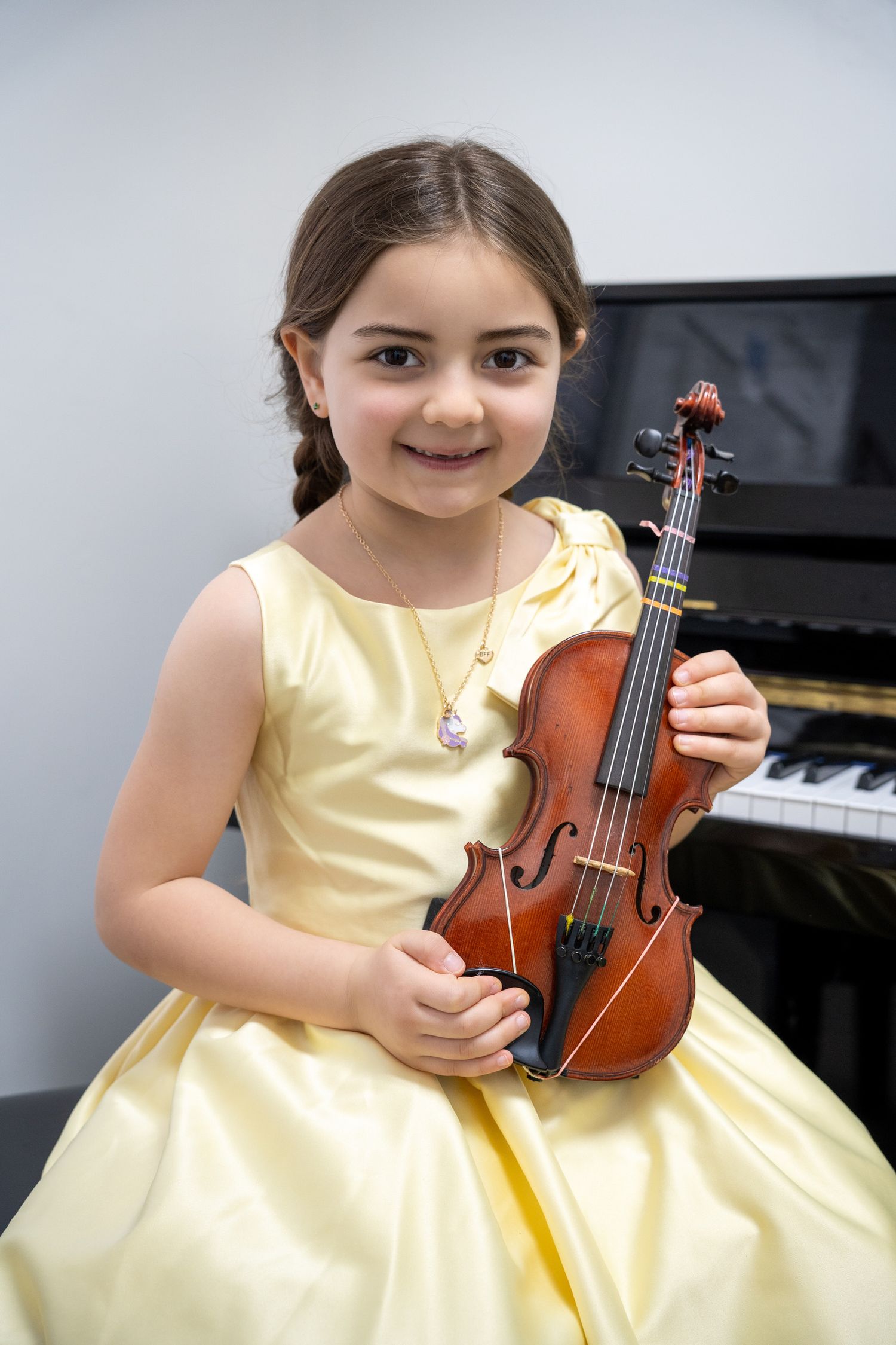 Portrait of a young girl in a yellow dress holding her violin in front of a piano.