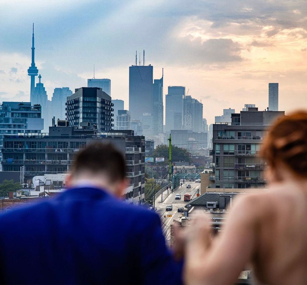 Two people enjoying a rooftop view of Toronto's skyline with the CN Tower visible during a hazy sunset.
