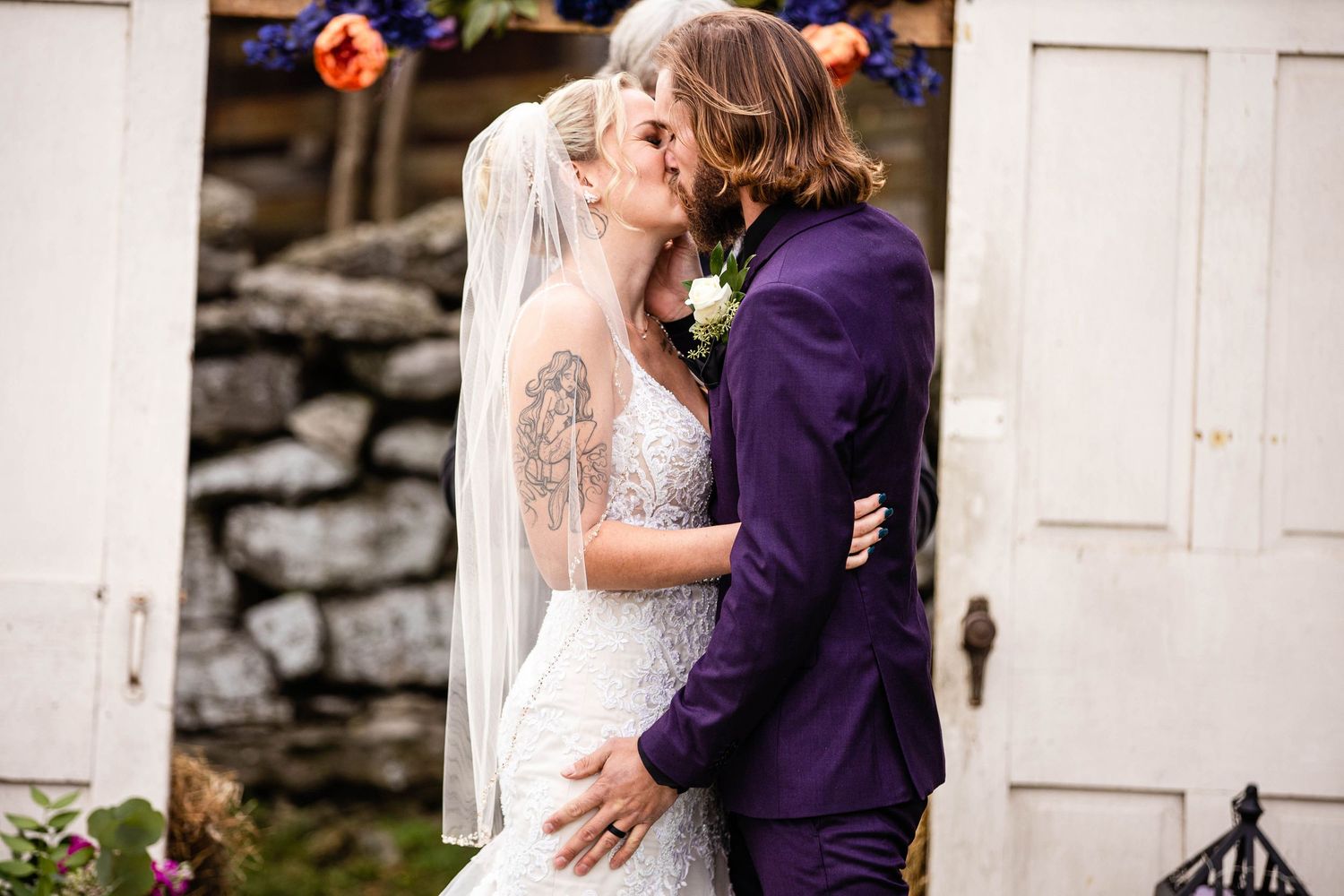A romantic wedding moment with the bride in white lace and groom in purple suit embracing and kissing outdoors.