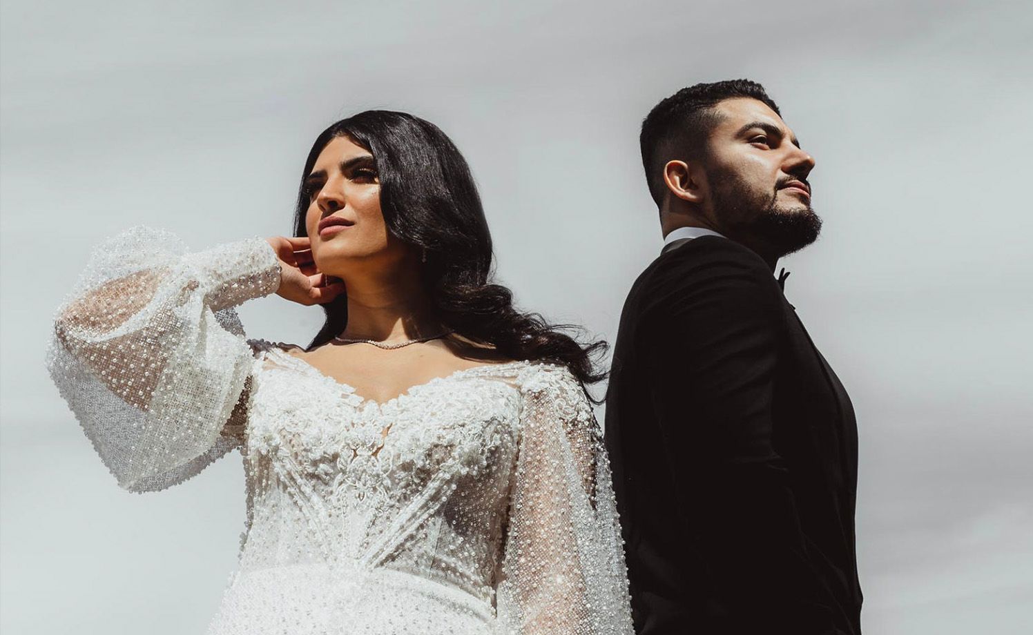 Dramatic side-by-side wedding portrait with bride in long-sleeve lace dress and groom in black suit against cloudy sky.