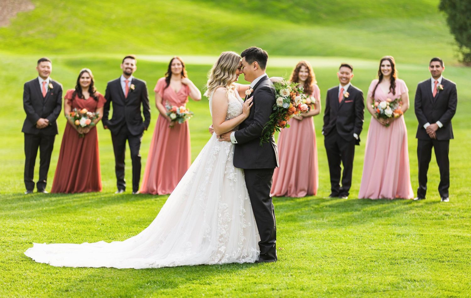 A couple shares their first kiss as newlyweds on a lush green hill while their wedding party looks on in pink and black attire.
