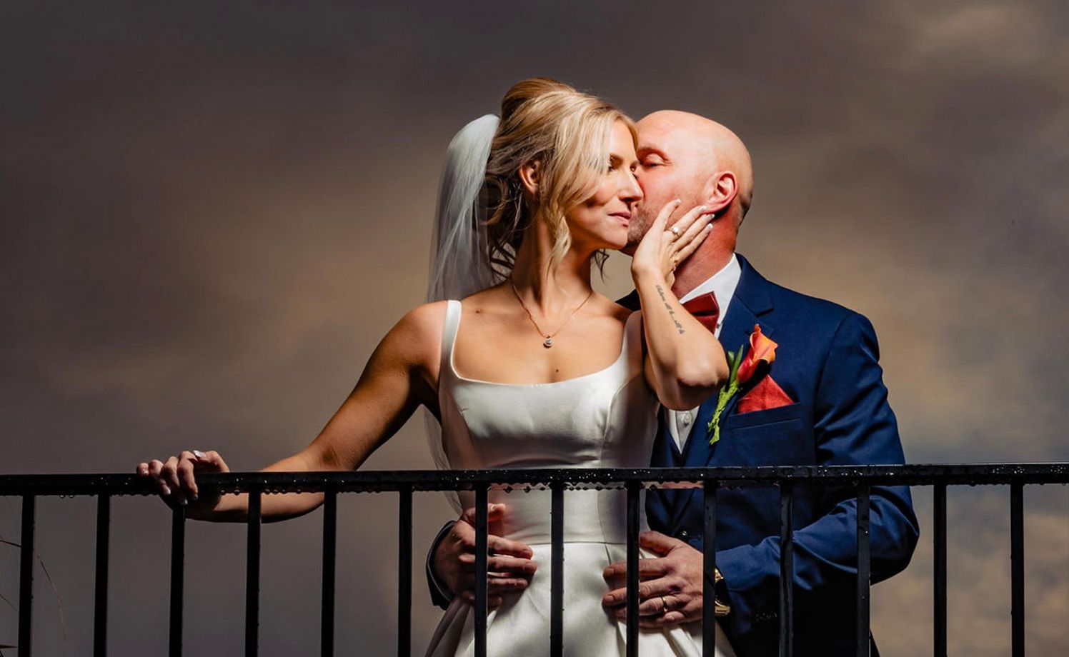 A joyful wedding couple shares a candid moment together while leaning on an outdoor railing at sunset.