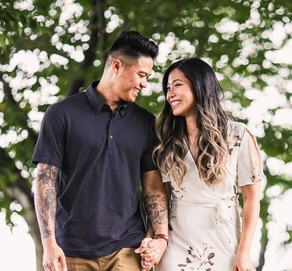 A smiling couple holding hands outdoors, with him in a navy polo and her in a floral cream dress.