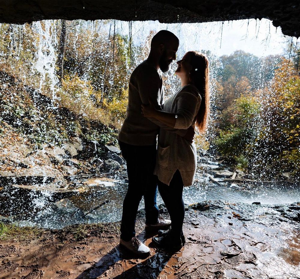 A silhouetted couple shares an intimate moment behind a flowing waterfall on a sunny autumn day.