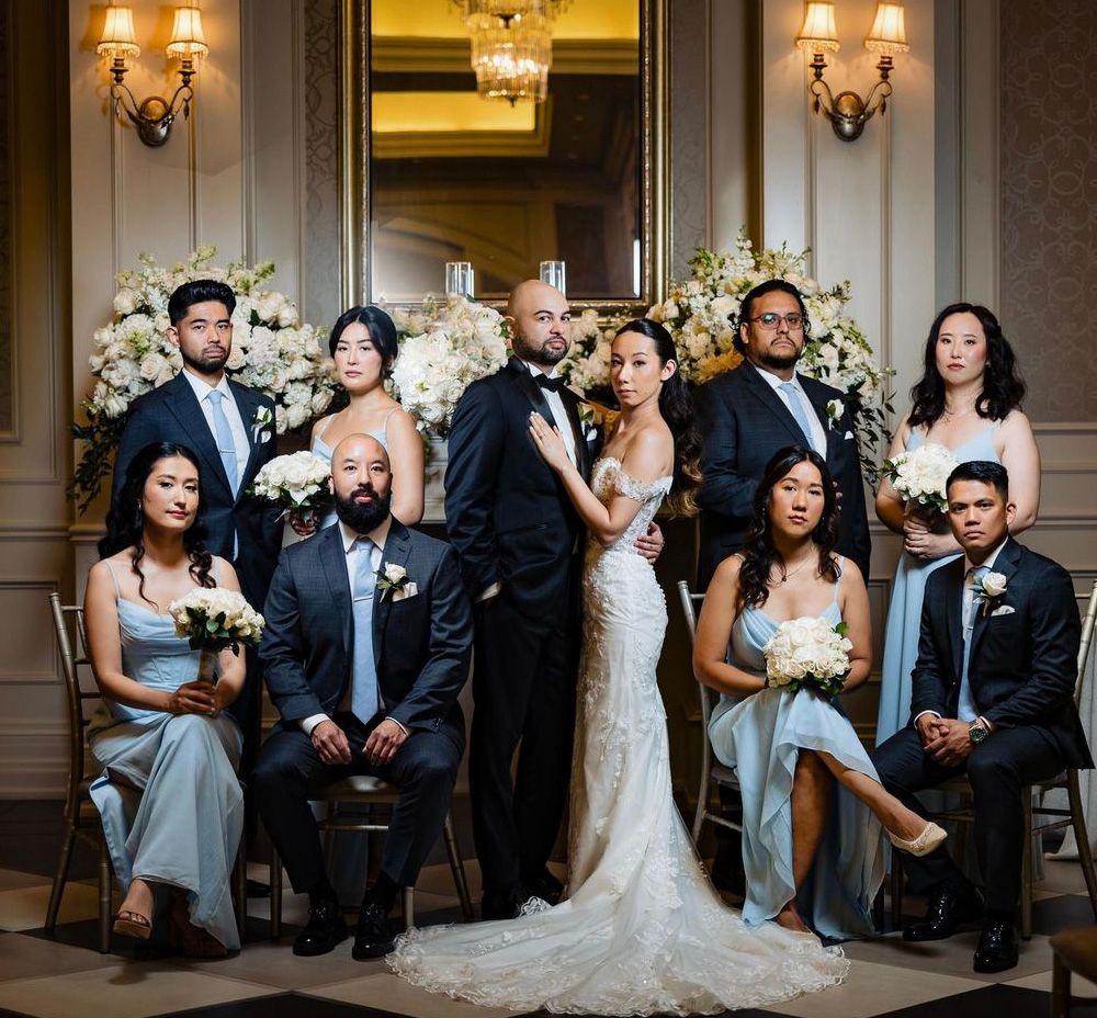 Elegant wedding party poses together in formal attire against ornate backdrop with floral arrangements and wall sconces.