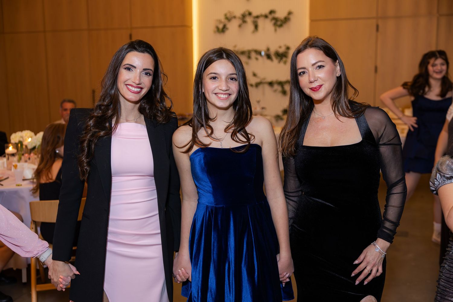 Three women in elegant dresses pose together at a formal event, smiling for the camera.