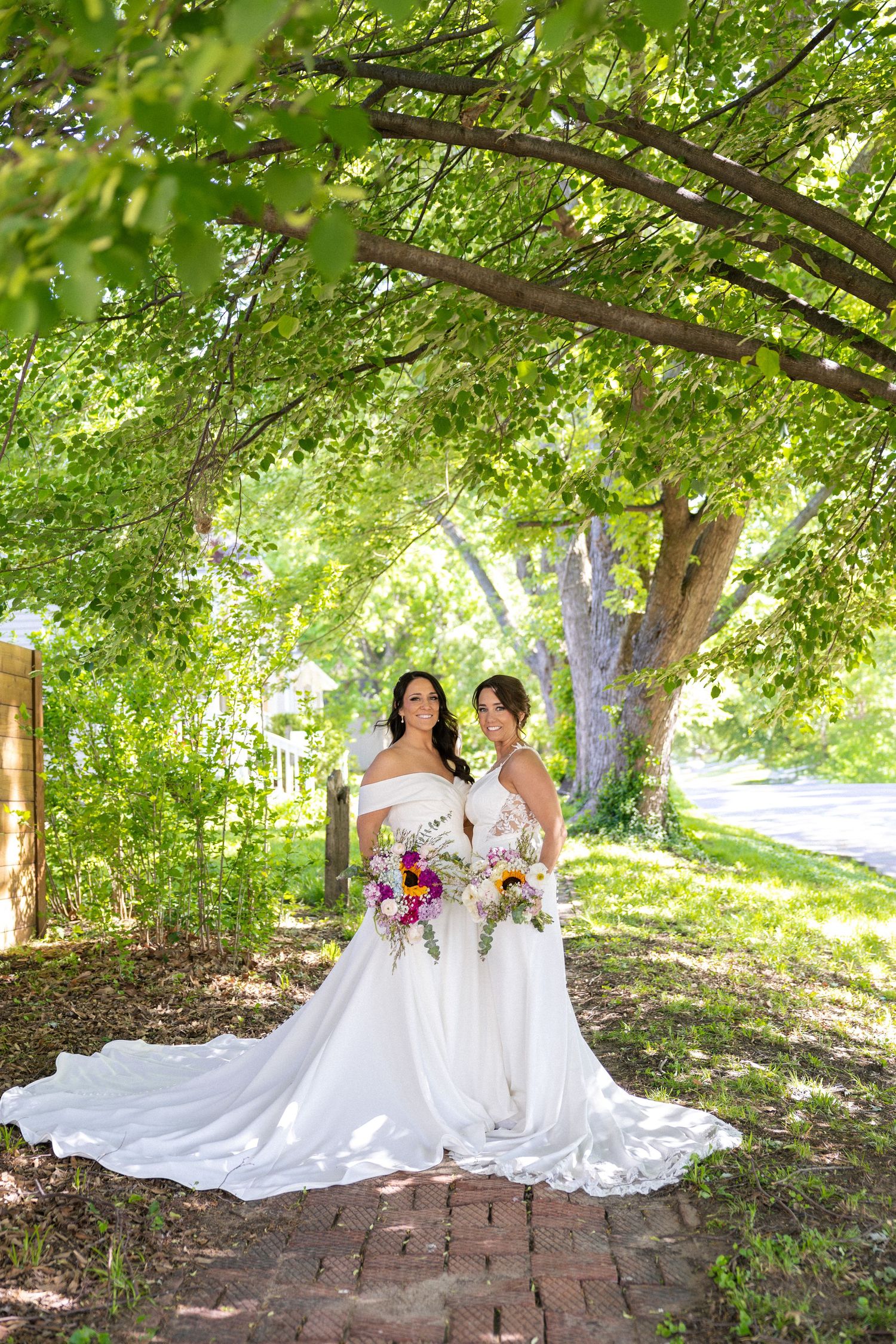 Two brides in elegant white gowns with bouquets standing under lush green tree canopy.