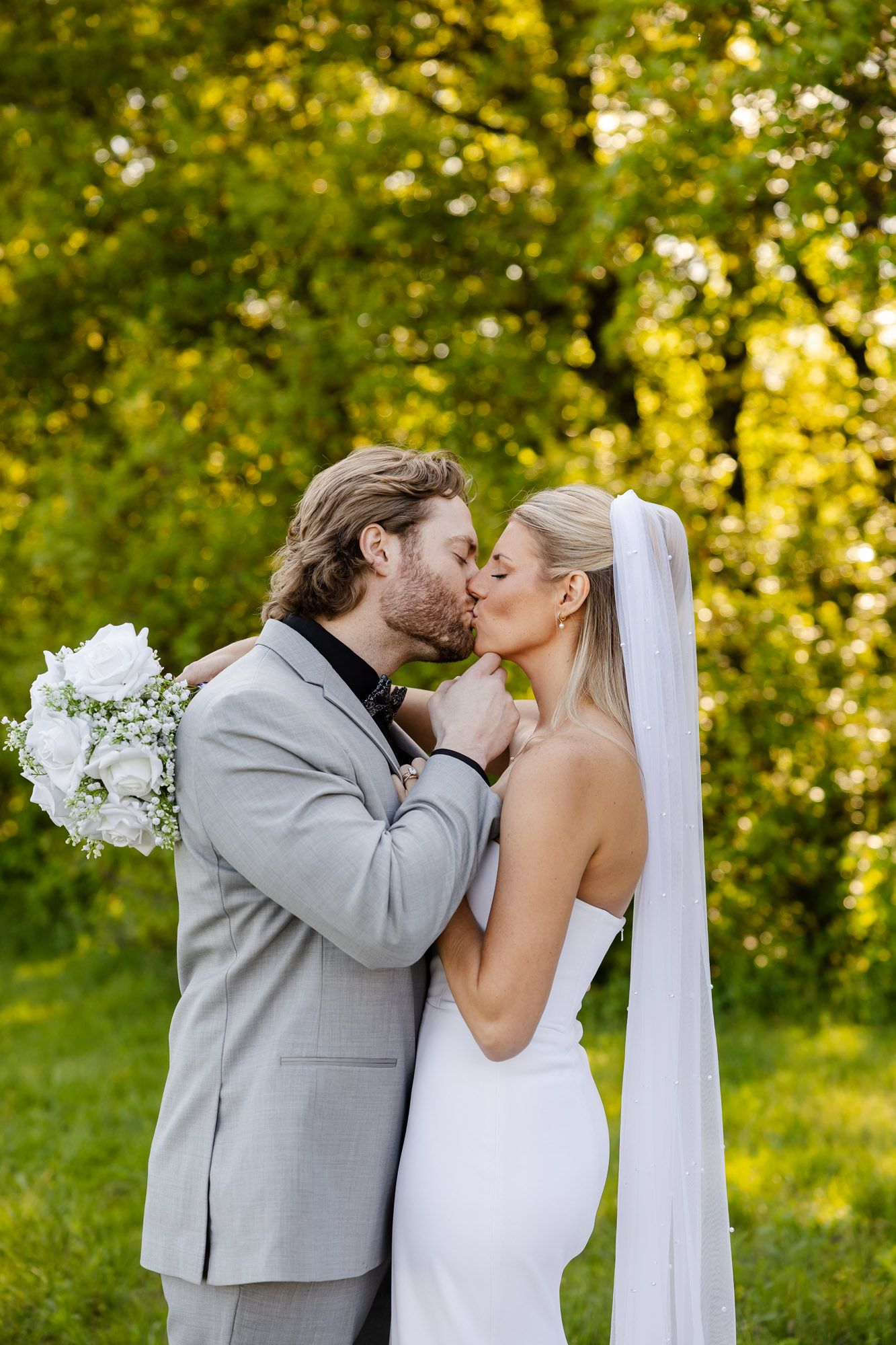 Bride and groom kissing outdoors surrounded by lush green trees on wedding day.