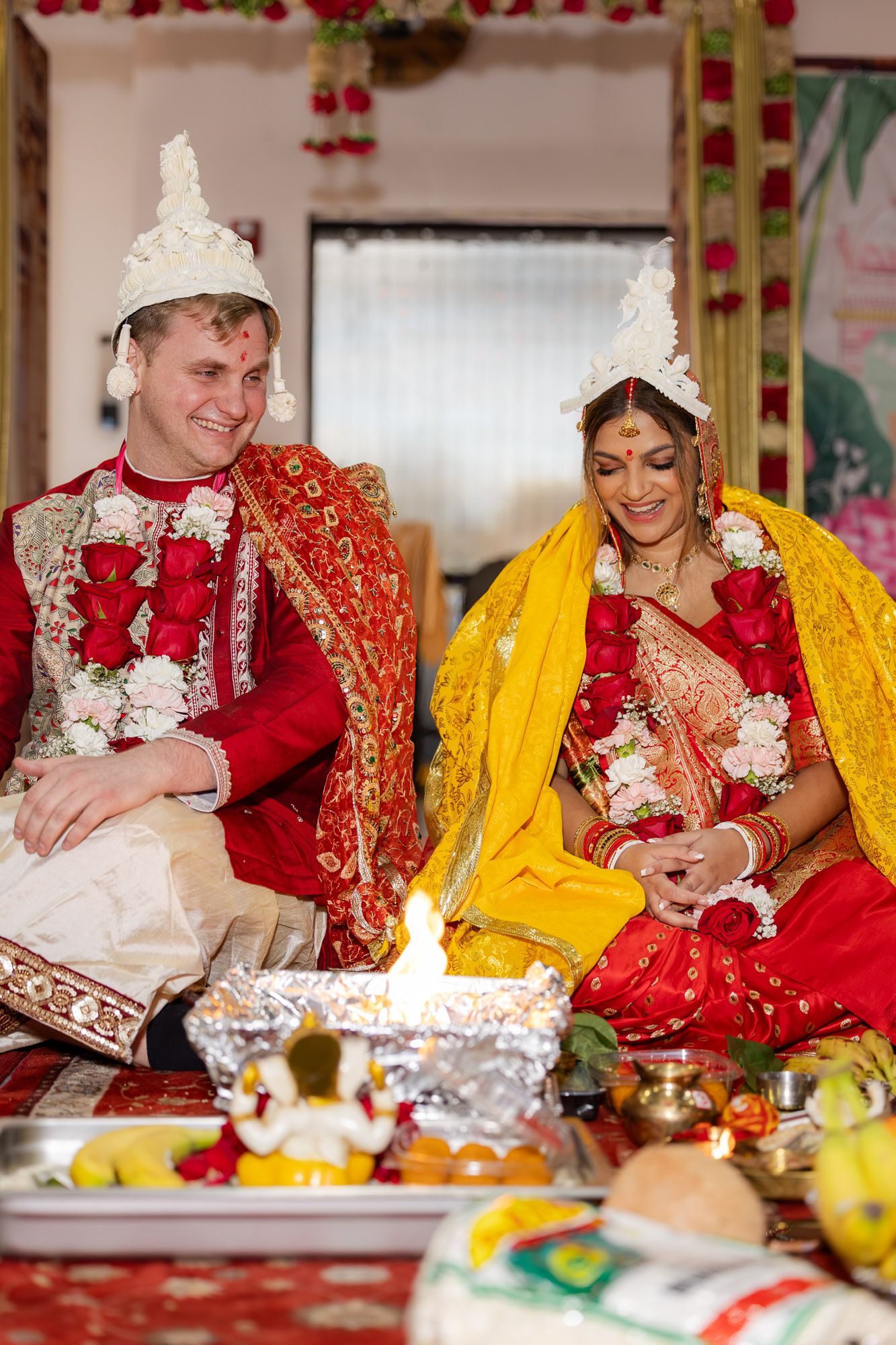 Couple in traditional Indian wedding attire smile during sacred fire ceremony ritual.