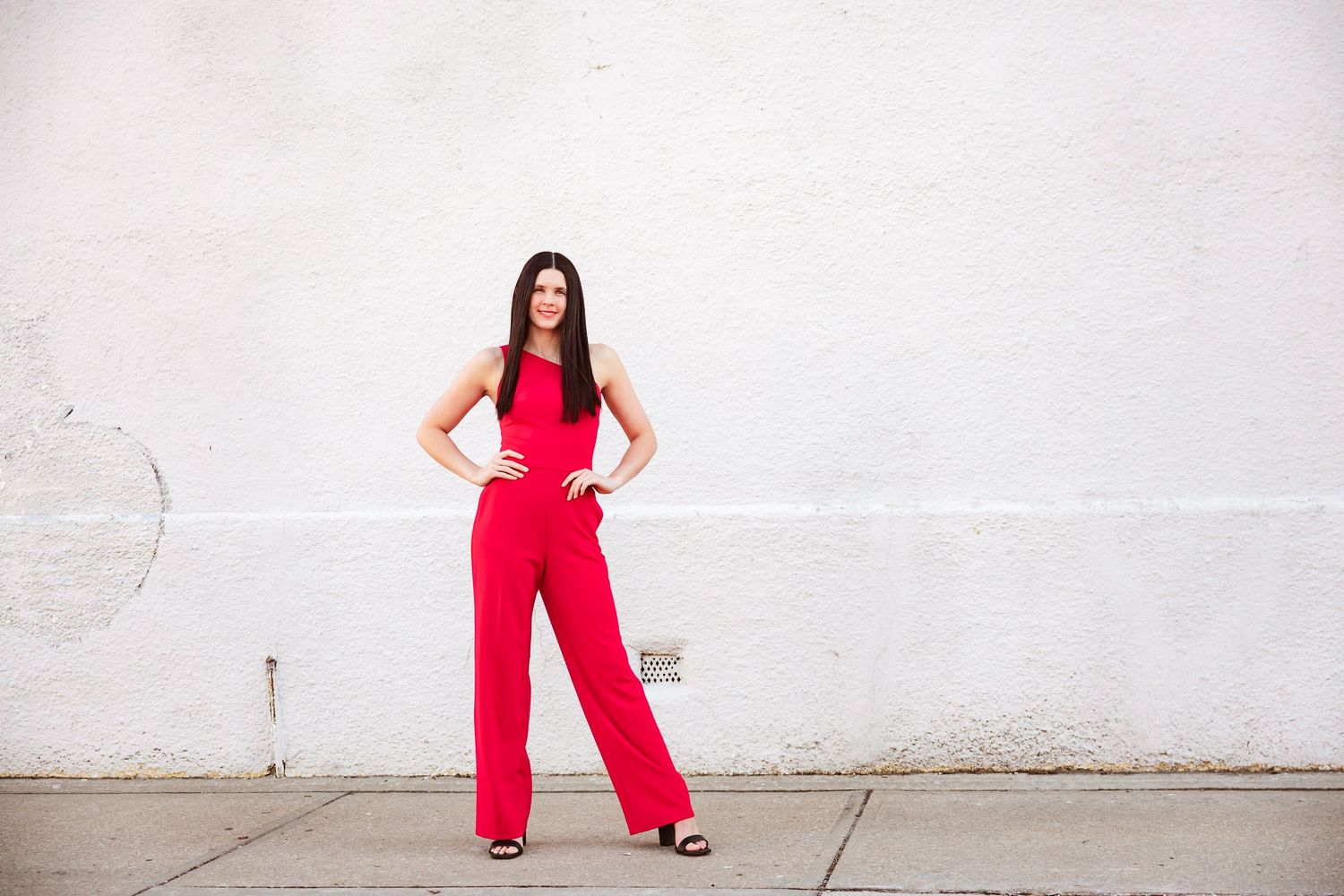 Confident woman posing in a red wide-leg jumpsuit against a white wall on a city sidewalk.