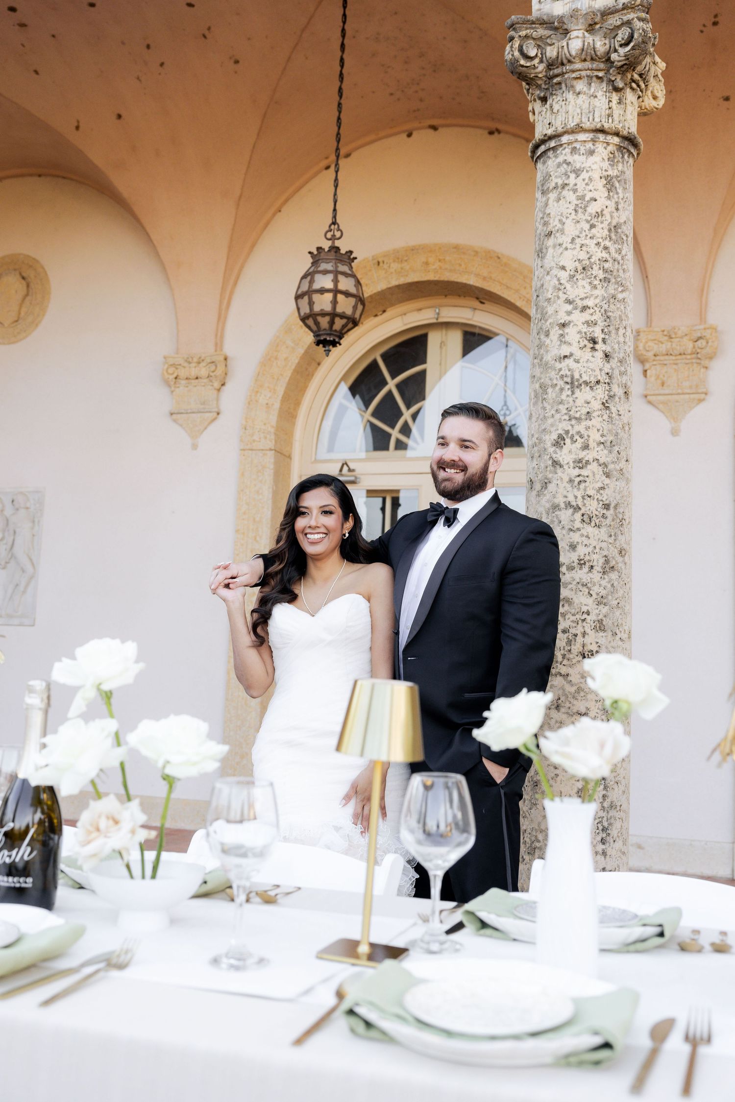 Smiling bride and groom stand at elegant white reception table with roses at historic venue.