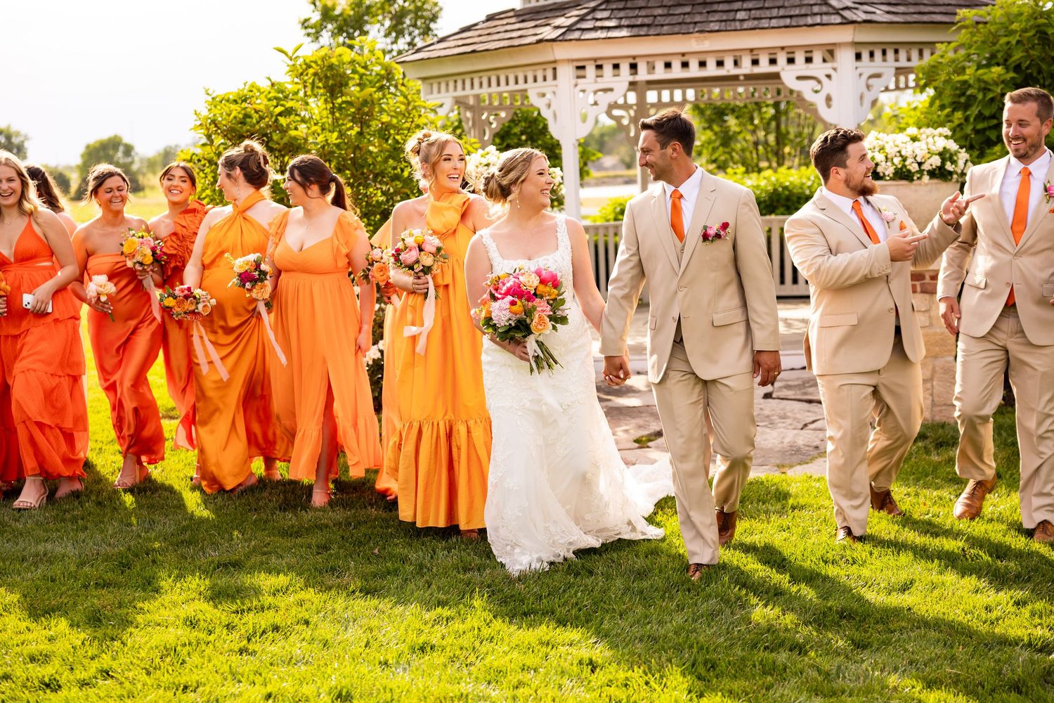 Wedding party walks together, bridesmaids in orange gowns and groomsmen in tan suits on sunny day.